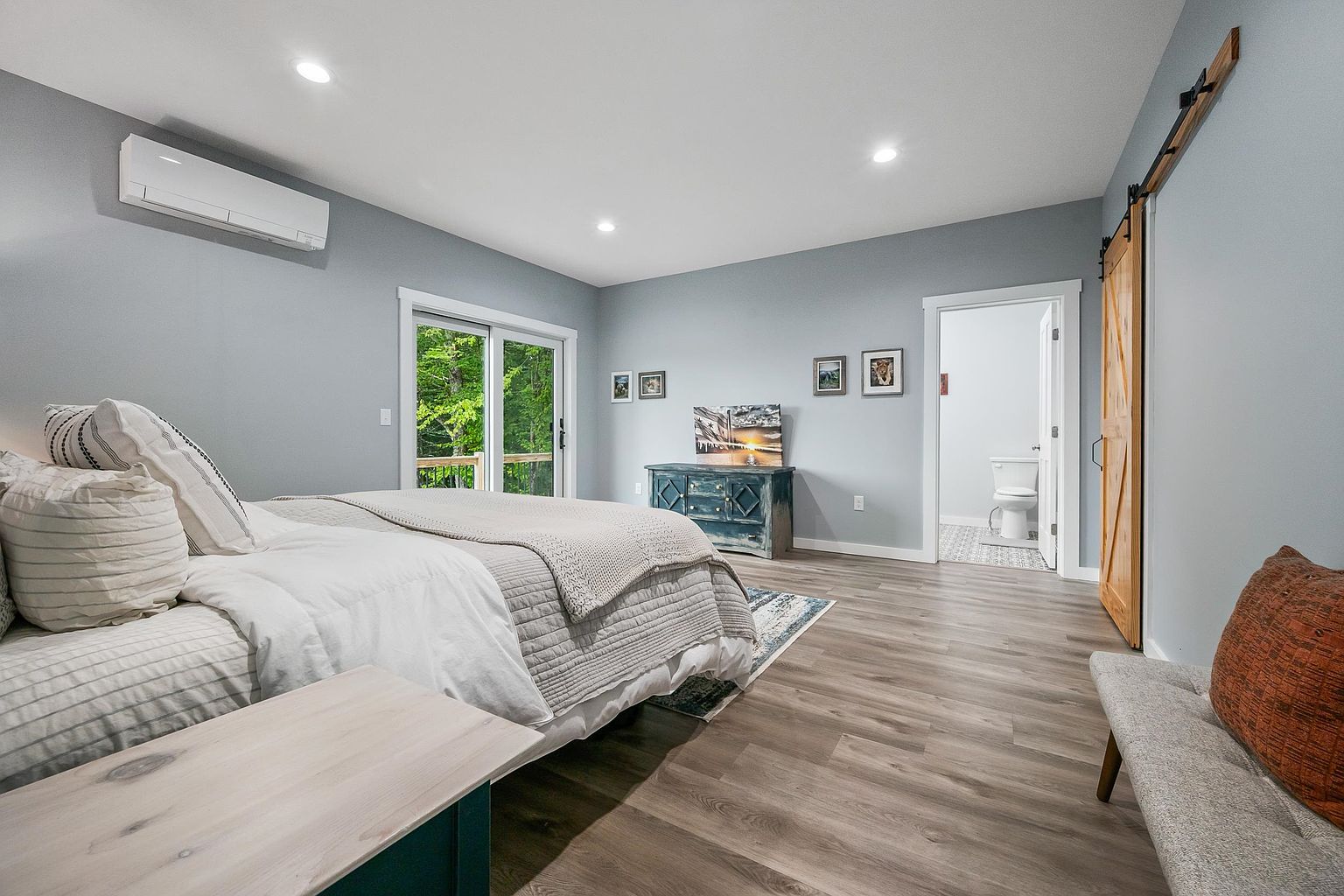 This spacious primary bedroom features light blue walls, wood-look flooring, and a large bed dressed in neutral linens. A sliding glass door provides access to an outdoor deck with a view of the surrounding trees, while a rustic wooden barn door leads to an attached bathroom. The room is accented by a distressed blue dresser, framed artwork, and a bench at the foot of the bed, creating a calm and inviting atmosphere.