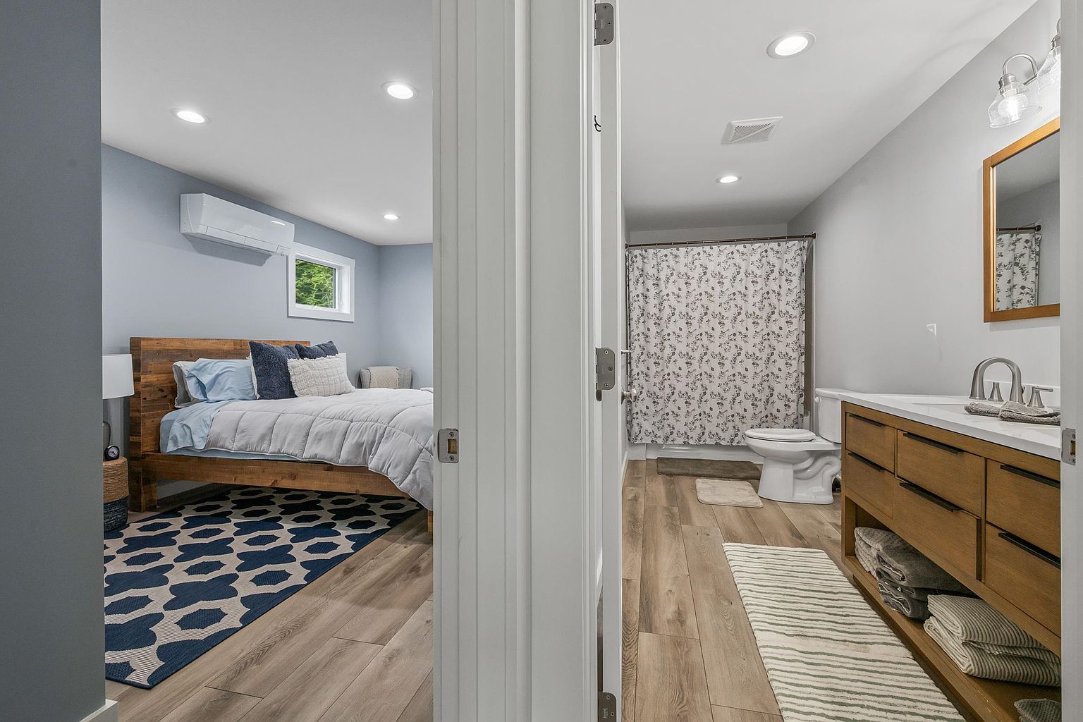 This perspective captures a transition space looking into both a bedroom and a bathroom, showcasing a cohesive design with light wood-look flooring throughout. The bedroom on the left features a rustic wood-frame bed and a bold geometric area rug, while the bathroom on the right includes a modern wood vanity and a floral-patterned shower curtain. The bright, neutral wall color and recessed lighting create an airy and inviting atmosphere.