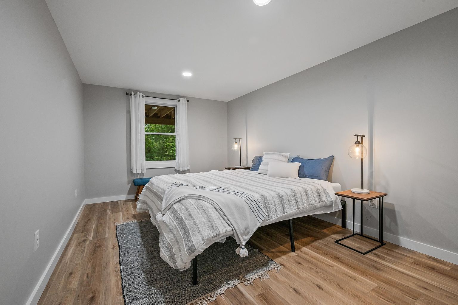 This bedroom features light-toned wood-look flooring and neutral gray walls, creating a clean and modern aesthetic. The room is furnished with a bed dressed in a striped comforter, flanked by two minimalist metal-frame side tables topped with globe-style lamps. A window with white curtains allows natural light to enter, and a small blue bench sits in the corner, providing a simple and inviting space.