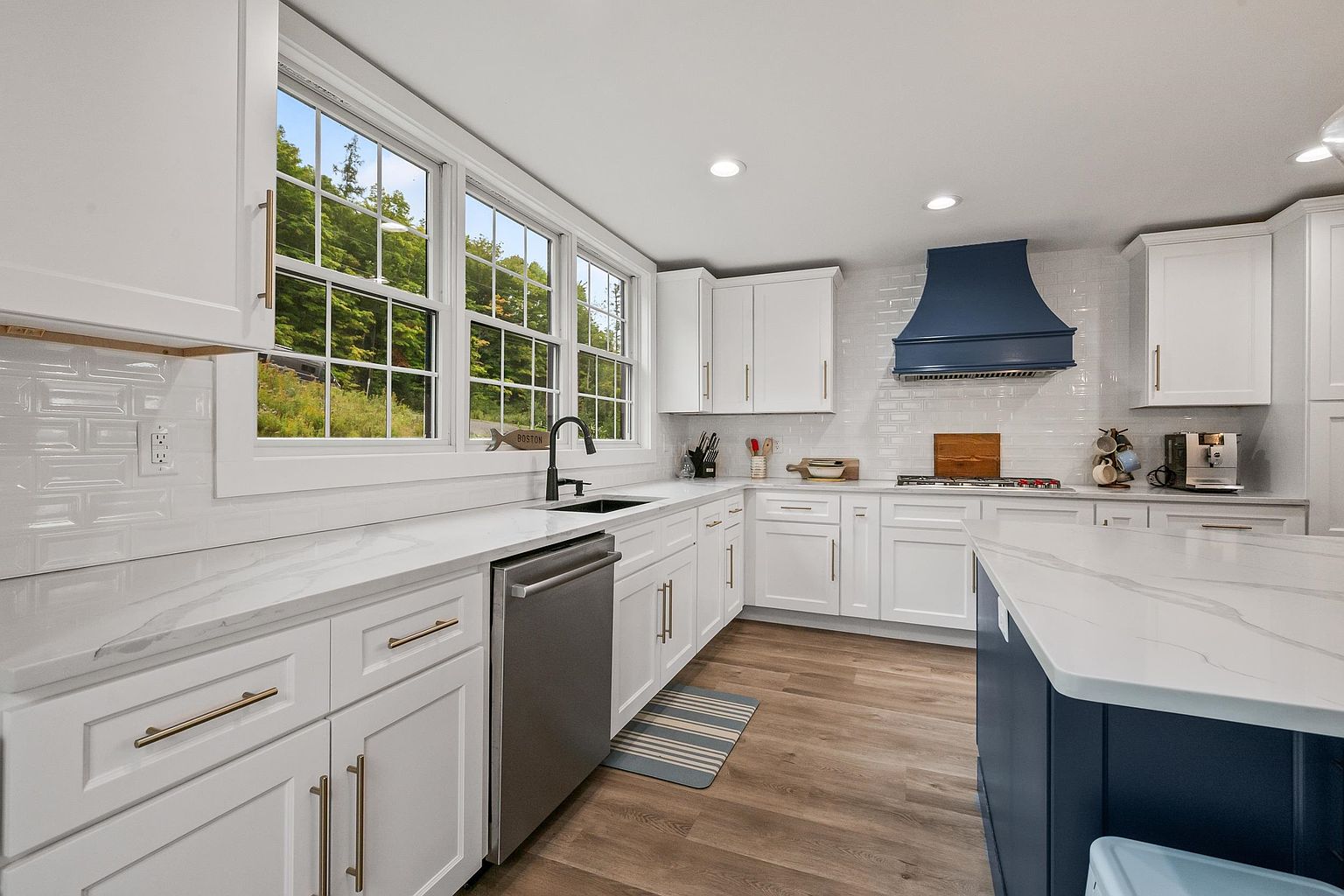 This bright and modern kitchen features crisp white cabinetry contrasted by a bold blue range hood and matching kitchen island base. The space is equipped with stainless steel appliances, a deep undermount sink, and light-toned stone countertops that complement the light wood-look flooring. Large windows above the sink provide ample natural light and a view of the surrounding greenery, creating an inviting and functional culinary space.