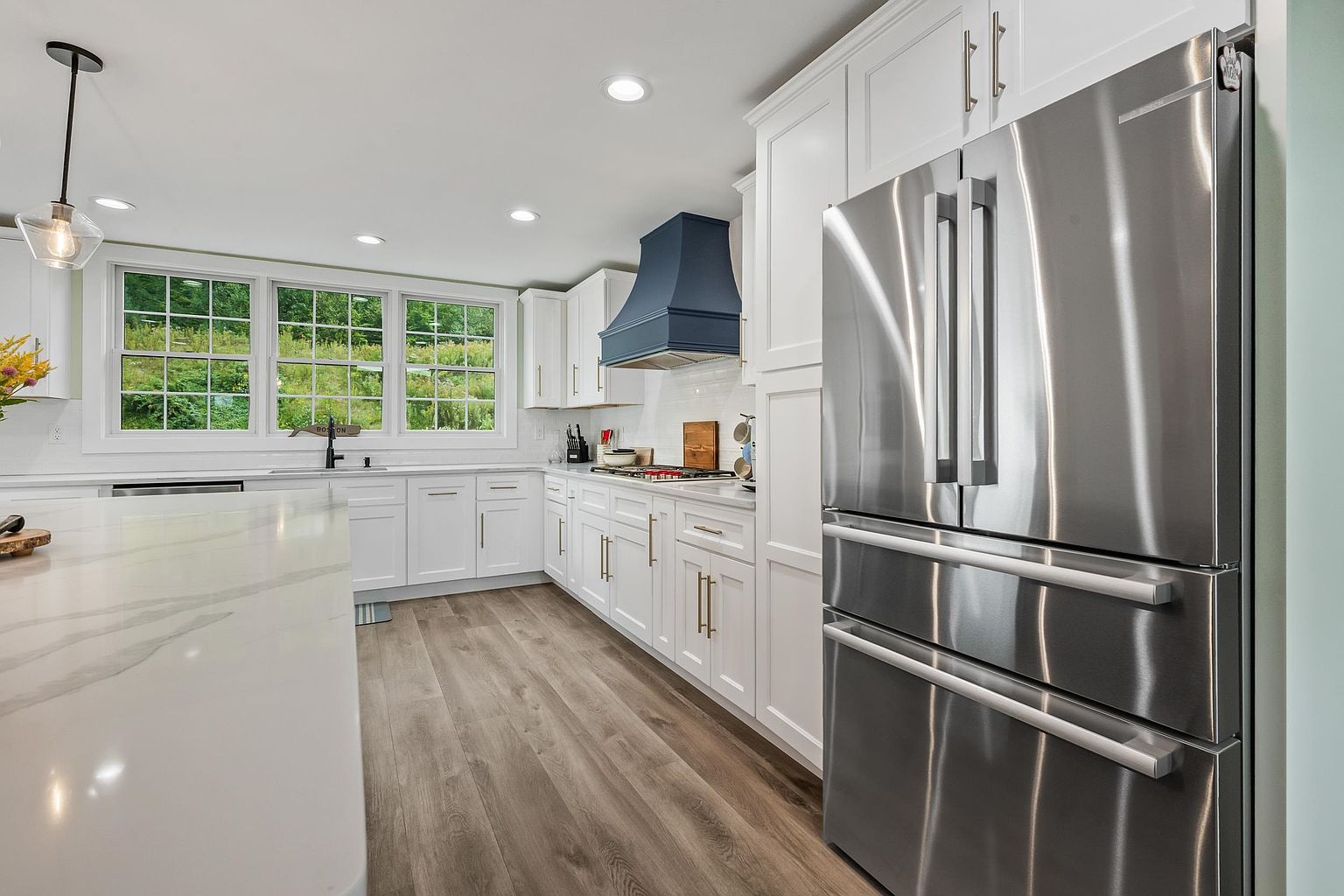This bright and modern kitchen features crisp white cabinetry, a large central island with a light-toned stone countertop, and high-end stainless steel appliances. A striking blue range hood serves as a focal point above the stove, while a row of windows above the sink provides ample natural light and a view of the outdoors. The space is finished with light wood-toned flooring, creating a clean, inviting, and contemporary atmosphere.