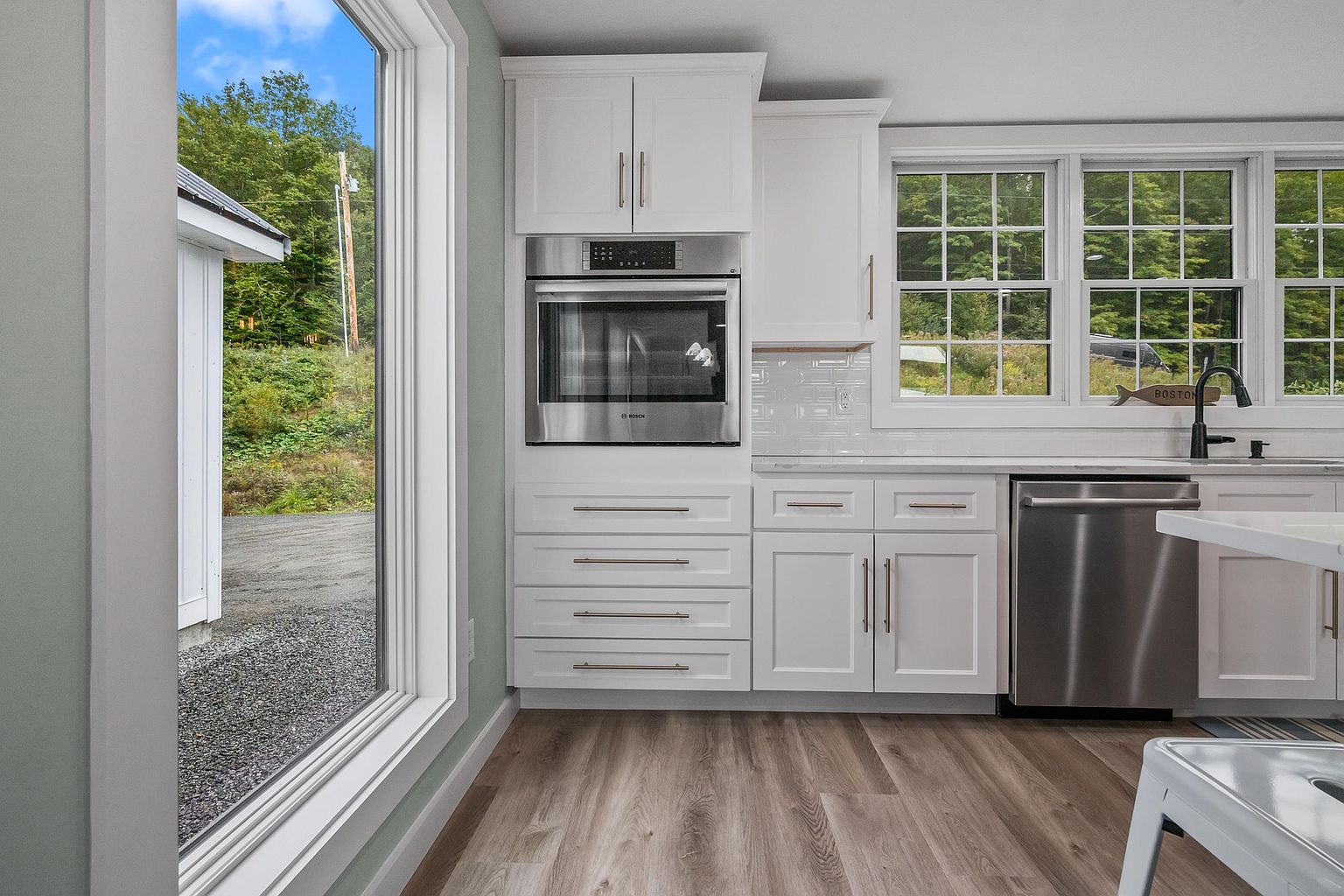 This bright, modern kitchen features crisp white cabinetry paired with stainless steel appliances, including a built-in oven and dishwasher. A large window provides a clear view of the exterior landscape, while a subway tile backsplash adds a clean, classic aesthetic. The space is finished with light-toned wood-look flooring, creating a cohesive and inviting atmosphere.