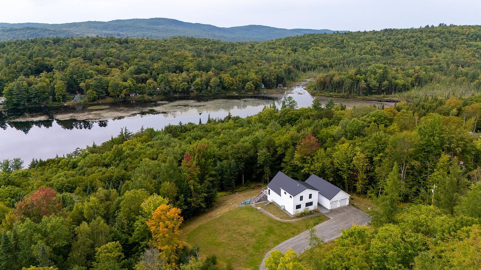This high-angle aerial shot captures a modern white house with a dark metal roof, situated on a lush, wooded lot overlooking a serene lake. The property features a paved driveway leading to an attached garage, a small grassy lawn, and a deck area, all surrounded by dense, mature forest. The perspective emphasizes the home's secluded, tranquil setting and its integration into the natural landscape.