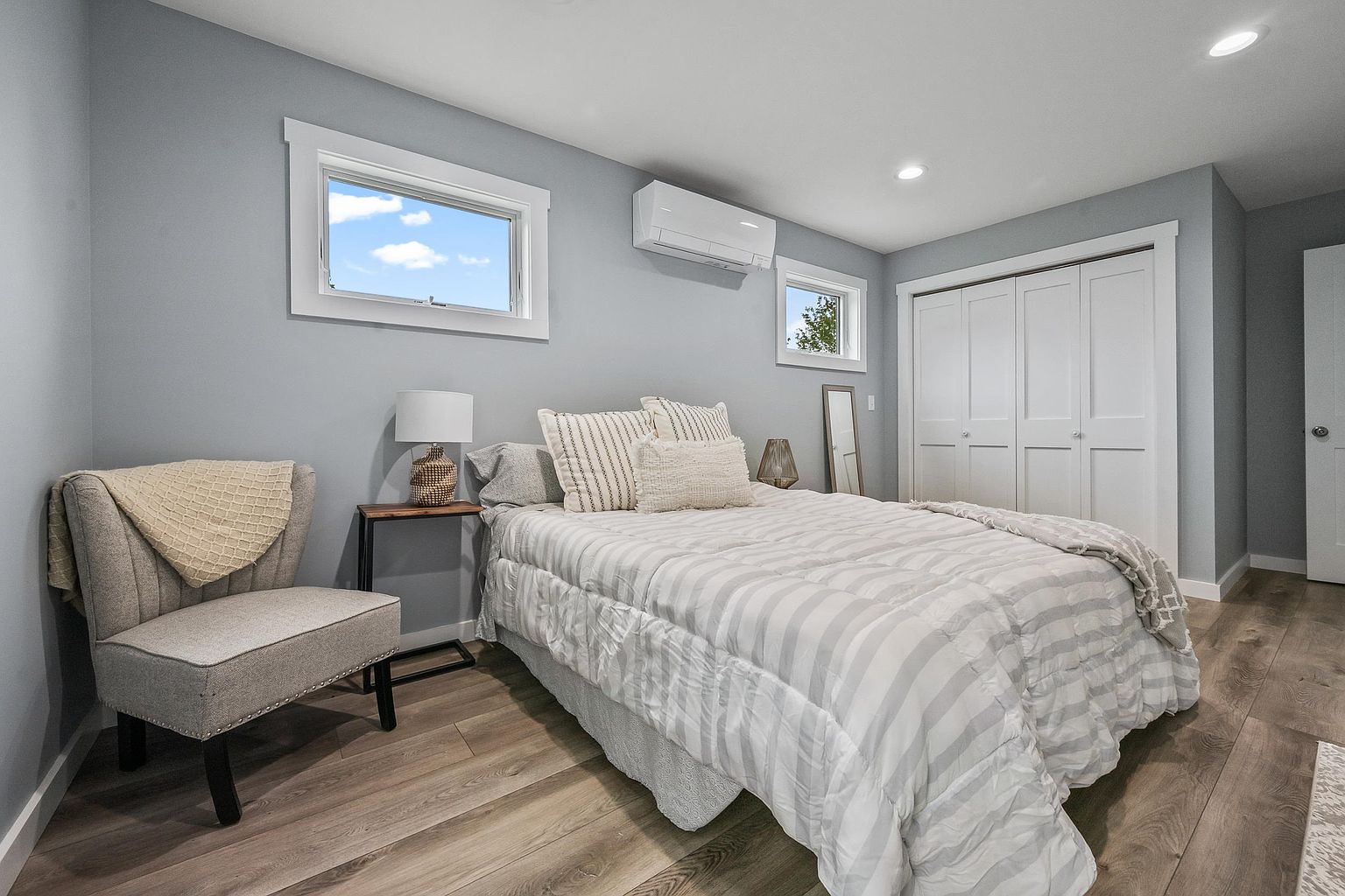 This bright and neutral bedroom features light gray walls, wood-look flooring, and a queen-sized bed dressed in a striped comforter. A cozy upholstered accent chair sits in the corner next to a slim side table, while a wall-mounted mini-split unit provides climate control. The room is illuminated by recessed ceiling lights and natural light streaming through two windows, creating a clean and inviting atmosphere.