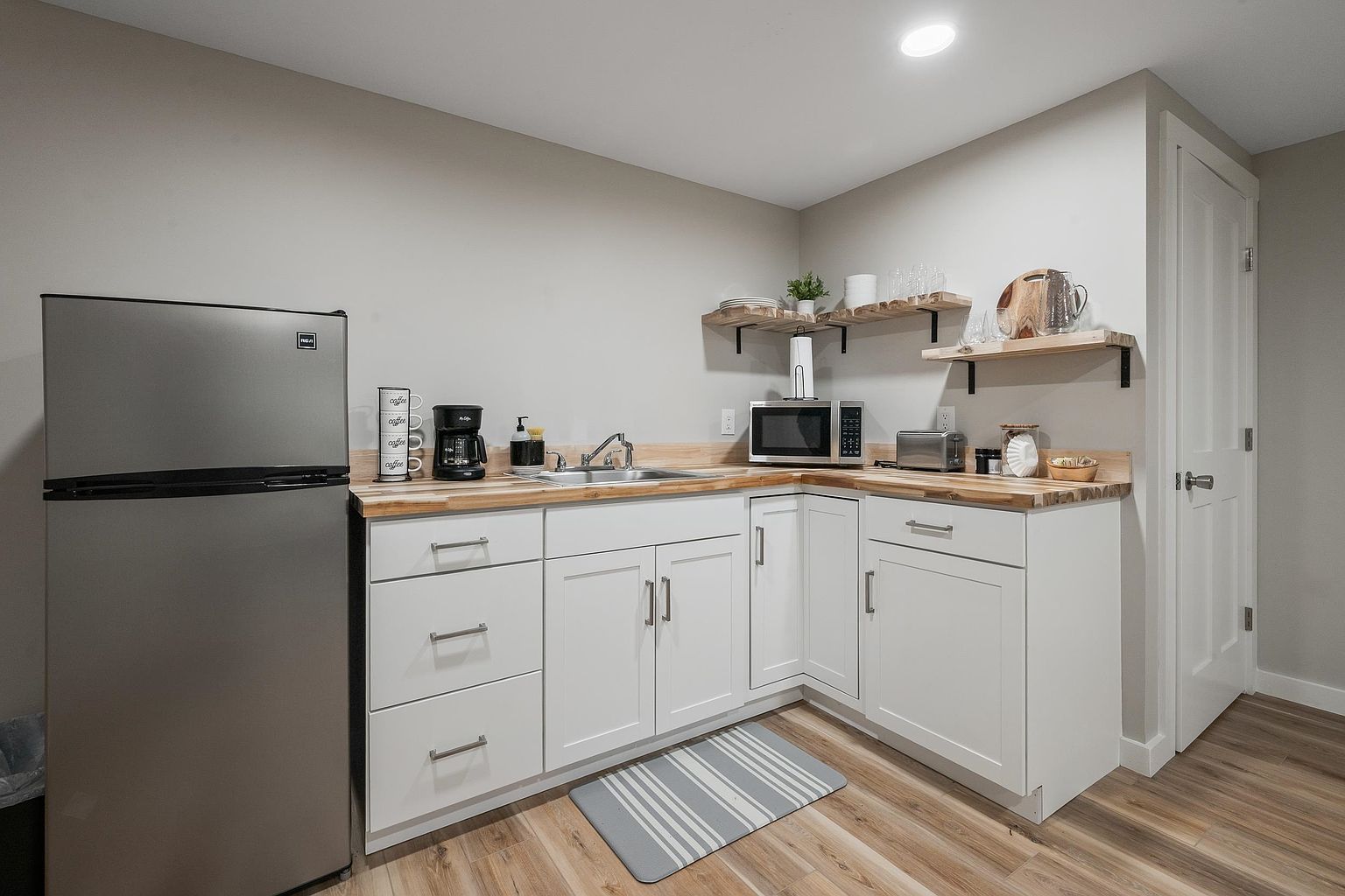 This compact kitchenette features white shaker-style cabinetry paired with a light-toned wood countertop and a stainless steel sink. The space is equipped with a stainless steel refrigerator, a microwave, and a coffee station, all set against a neutral wall color. Floating wood shelves provide open storage, while a striped rug adds a touch of texture to the wood-look flooring.