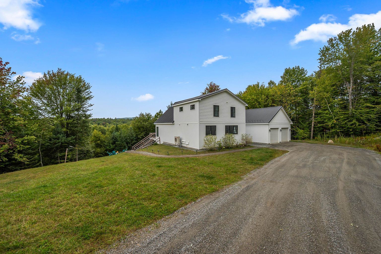 This side view of a modern, white-sided home showcases a clean, minimalist aesthetic with a dark metal roof and a two-car attached garage. A gravel driveway leads up to the structure, which is situated on a gentle, grassy slope surrounded by lush, mature trees. The perspective captures the home's integration into the natural landscape, highlighting the peaceful, rural setting.