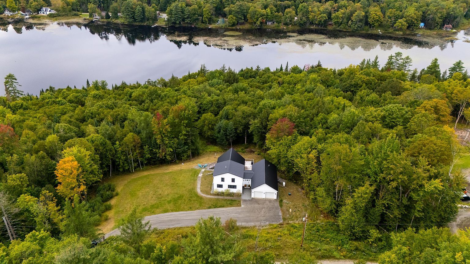 This high-angle aerial view captures a modern white house with a dark metal roof nestled within a lush, forested landscape. The property features a paved driveway leading to an attached garage, a spacious grassy yard, and a serene lake visible in the background. The setting offers a peaceful, secluded atmosphere surrounded by dense greenery.