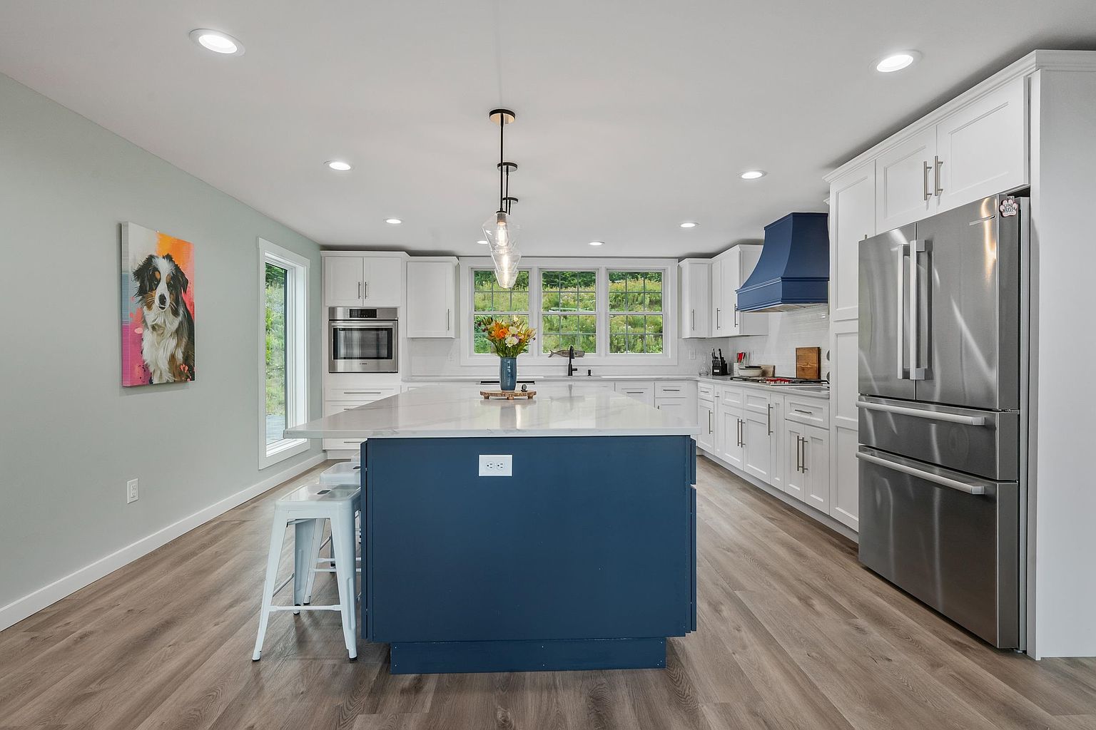 This bright and modern kitchen features a large central island with a deep blue base and a light-toned countertop, complemented by white cabinetry and a matching blue range hood. The space is illuminated by recessed lighting and a stylish pendant fixture, while light wood-look flooring runs throughout. A large window above the sink provides a view of the outdoors, and a colorful dog portrait adds a personalized touch to the light green wall.