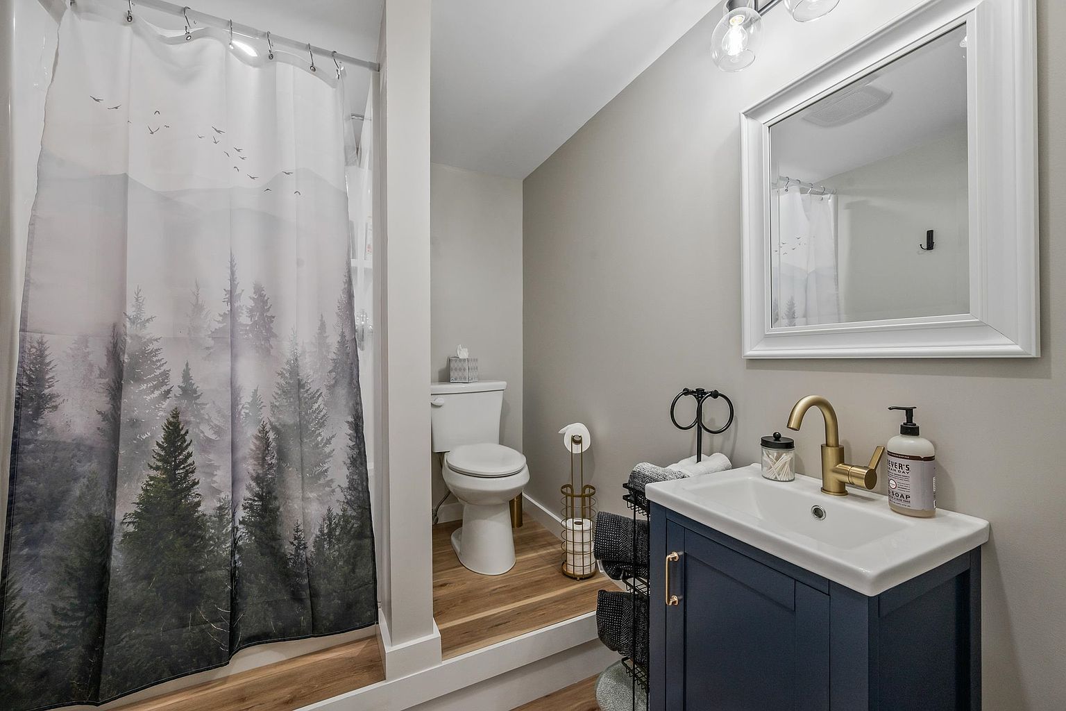 This compact bathroom features a raised platform area for the toilet, accented by wood-look flooring and a nature-themed shower curtain. A navy blue vanity with a gold faucet and a white rectangular mirror creates a modern, stylish contrast against the neutral gray walls. The space is well-lit by a multi-bulb vanity light, offering a clean and functional design.