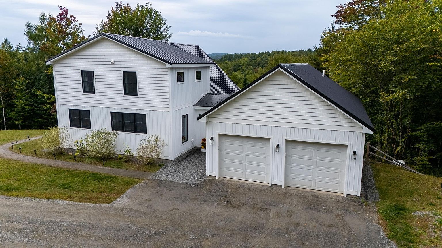 This elevated perspective captures a modern farmhouse-style home featuring crisp white vertical siding and a dark metal roof. The property includes an attached two-car garage and is set against a backdrop of lush, dense forest. A gravel driveway leads to the garage, while a paved walkway winds through the lawn, emphasizing the home's serene and secluded rural setting.