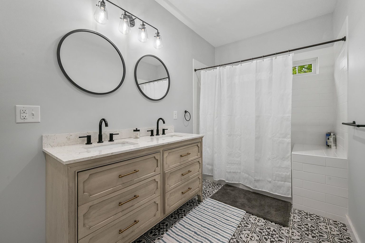 This bright bathroom features a double vanity with a light wood finish, topped with a stone counter and dual black faucets. Above the vanity, two circular mirrors are mounted on a light gray wall, illuminated by a four-light industrial-style fixture. The space includes a shower-tub combination with a white curtain and patterned tile flooring, creating a clean and modern aesthetic.