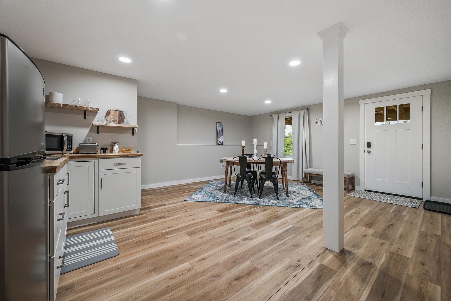 This open-concept space features a functional kitchen area with white cabinetry, light wood-toned countertops, and stainless steel appliances, seamlessly transitioning into a dining area. The room is anchored by a large, patterned area rug under a wooden dining table with black metal chairs, all set against neutral grey walls and light wood-look flooring. A prominent white structural column stands in the foreground, while a white door with glass panes provides natural light and entry to the space.