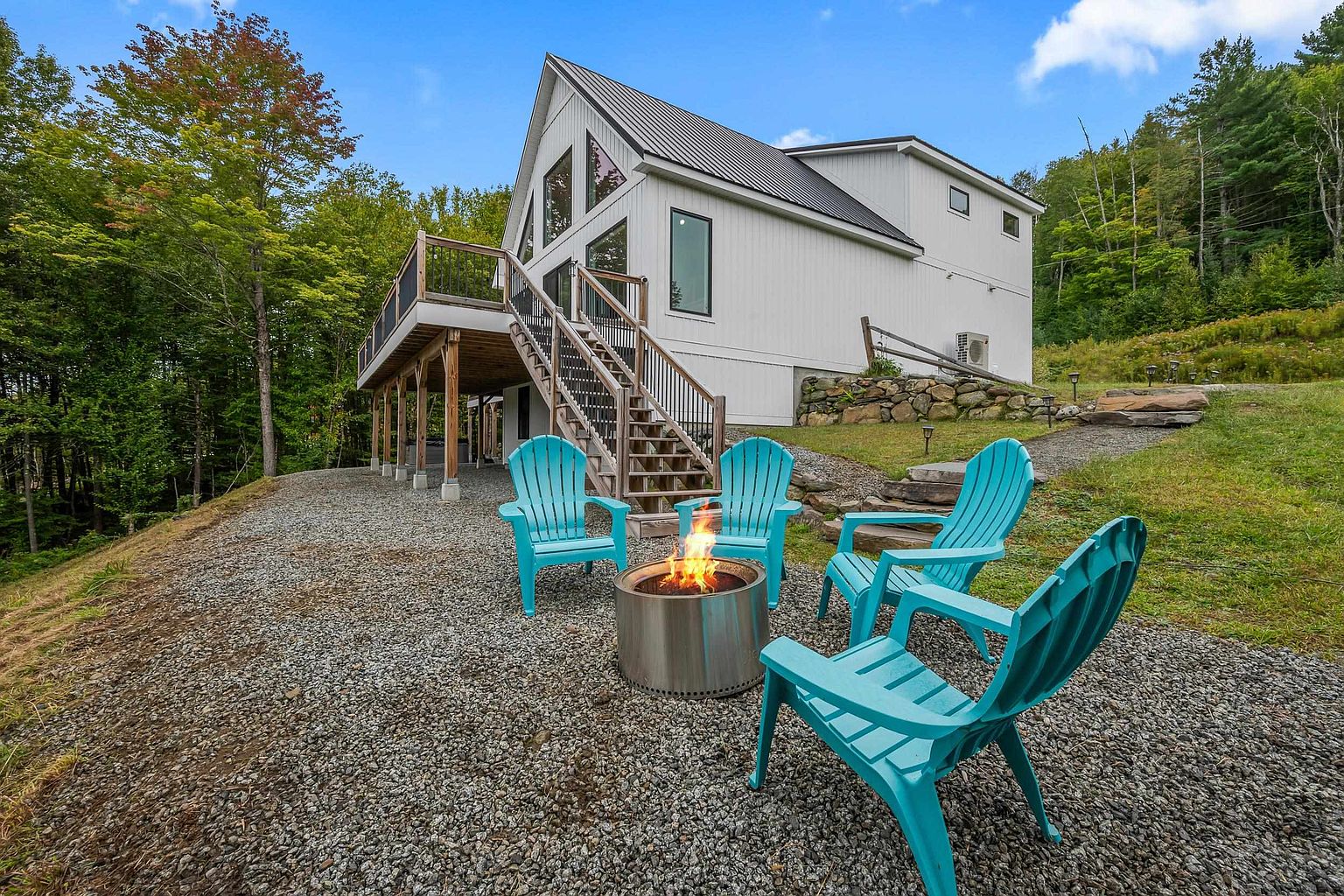 This rear view of a modern, light-colored home showcases a spacious elevated deck with wooden supports and a metal railing. Below the deck, a gravel patio area features a circular metal fire pit surrounded by four bright teal Adirondack chairs, creating an inviting outdoor living space. The property is nestled against a lush, wooded backdrop, emphasizing a peaceful and private setting.