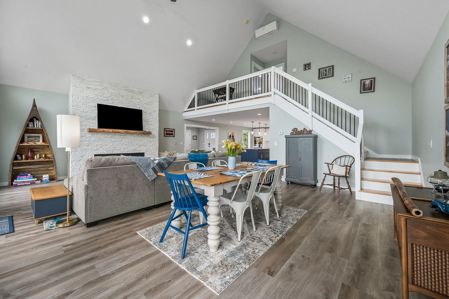 This open-concept living and dining area features a vaulted ceiling and a prominent stone-clad chimney breast with a wooden mantel. A cozy gray sectional sofa faces the fireplace, while a dining table with a mix of blue and white chairs sits on a patterned rug in the foreground. The space includes a lofted area with a white railing overlooking the main floor, creating an airy, multi-level atmosphere with light-toned wood-look flooring throughout.