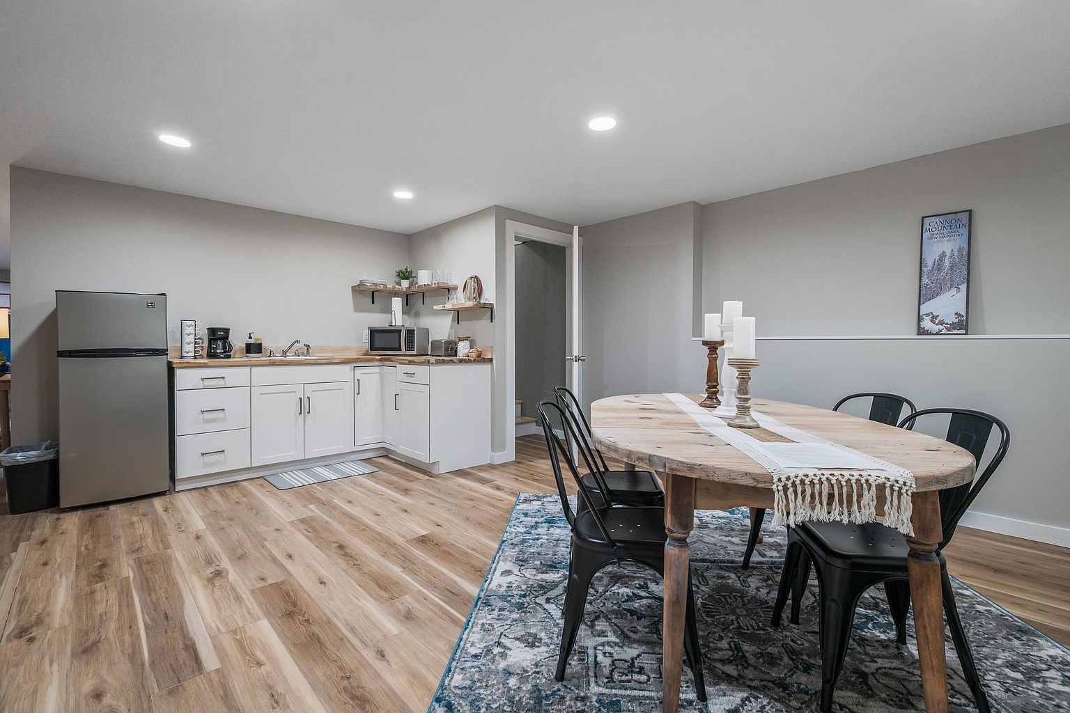 This open-concept space features a functional kitchenette with white cabinetry, a stainless steel refrigerator, and light wood-toned countertops. Adjacent to the kitchen area is a dining space anchored by an oval wooden table surrounded by black metal chairs, all set upon a large, patterned area rug. The room is finished with light-colored walls and wood-look flooring, creating a bright and inviting atmosphere suitable for casual dining or entertaining.