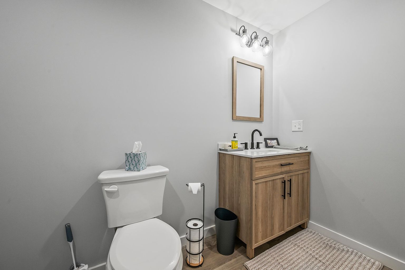 This compact powder room features a light wood vanity with a white countertop and a modern dark faucet. A simple rectangular mirror hangs above the vanity, illuminated by a three-light fixture, while a white toilet and a freestanding toilet paper holder occupy the adjacent space. The room is finished with neutral gray walls and light-toned flooring, creating a clean and functional aesthetic.