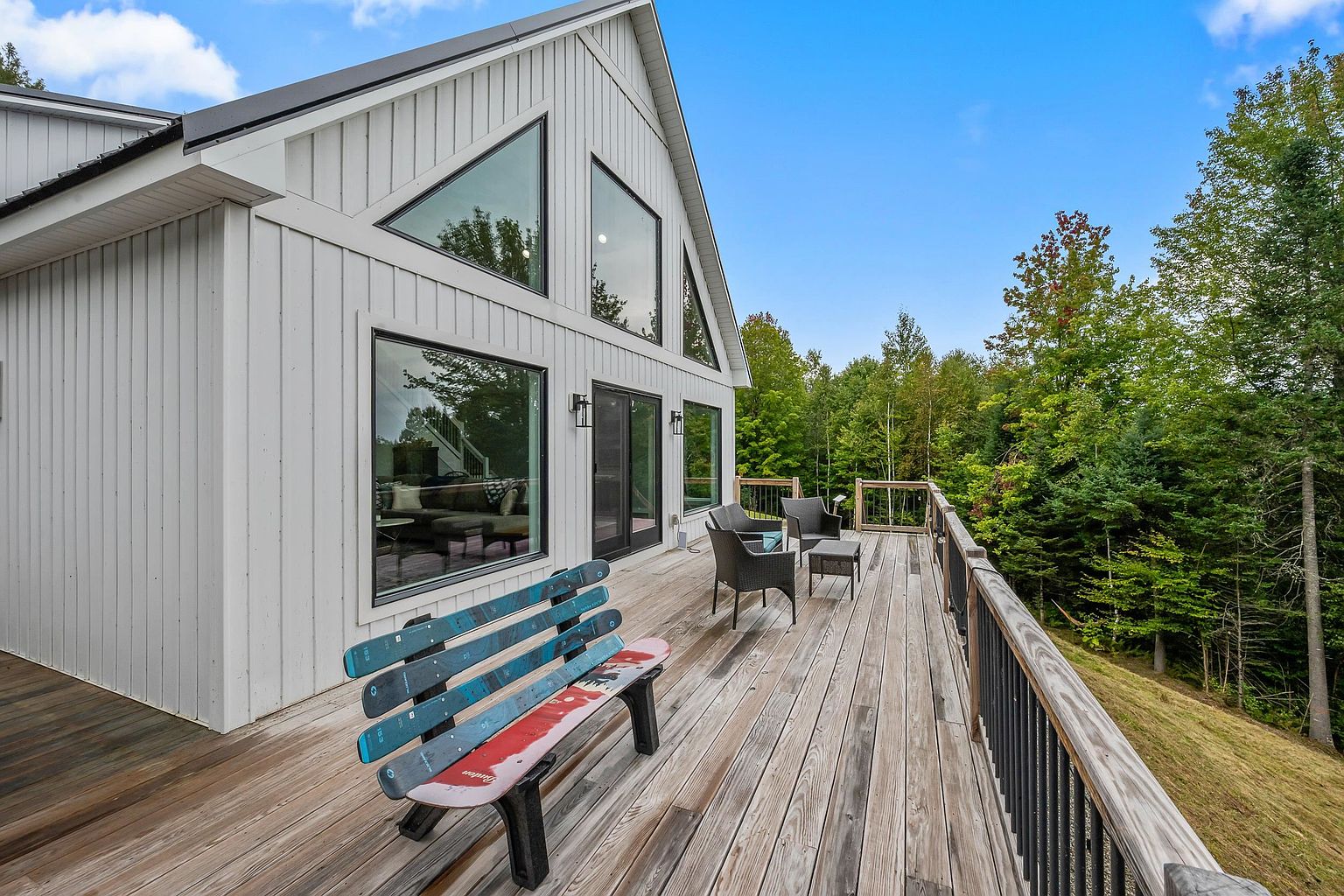 This expansive wooden deck offers a scenic outdoor living space, featuring a unique bench crafted from repurposed skis and a small seating area with dark wicker furniture. The home showcases a modern farmhouse aesthetic with white vertical siding, black-framed windows, and a steep roofline. The deck overlooks a lush, tree-filled landscape, providing a serene and elevated perspective of the surrounding natural environment.