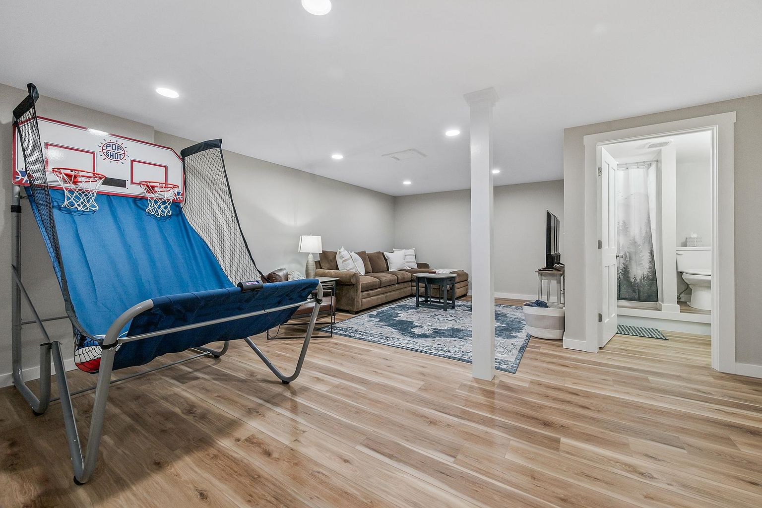 This finished basement features light wood-look flooring and a neutral color palette, creating a versatile living space. The area includes a cozy seating arrangement with a brown sectional sofa and a patterned area rug, alongside a basketball arcade game in the foreground. A doorway to the right leads into a bathroom, and the room is illuminated by recessed lighting throughout.