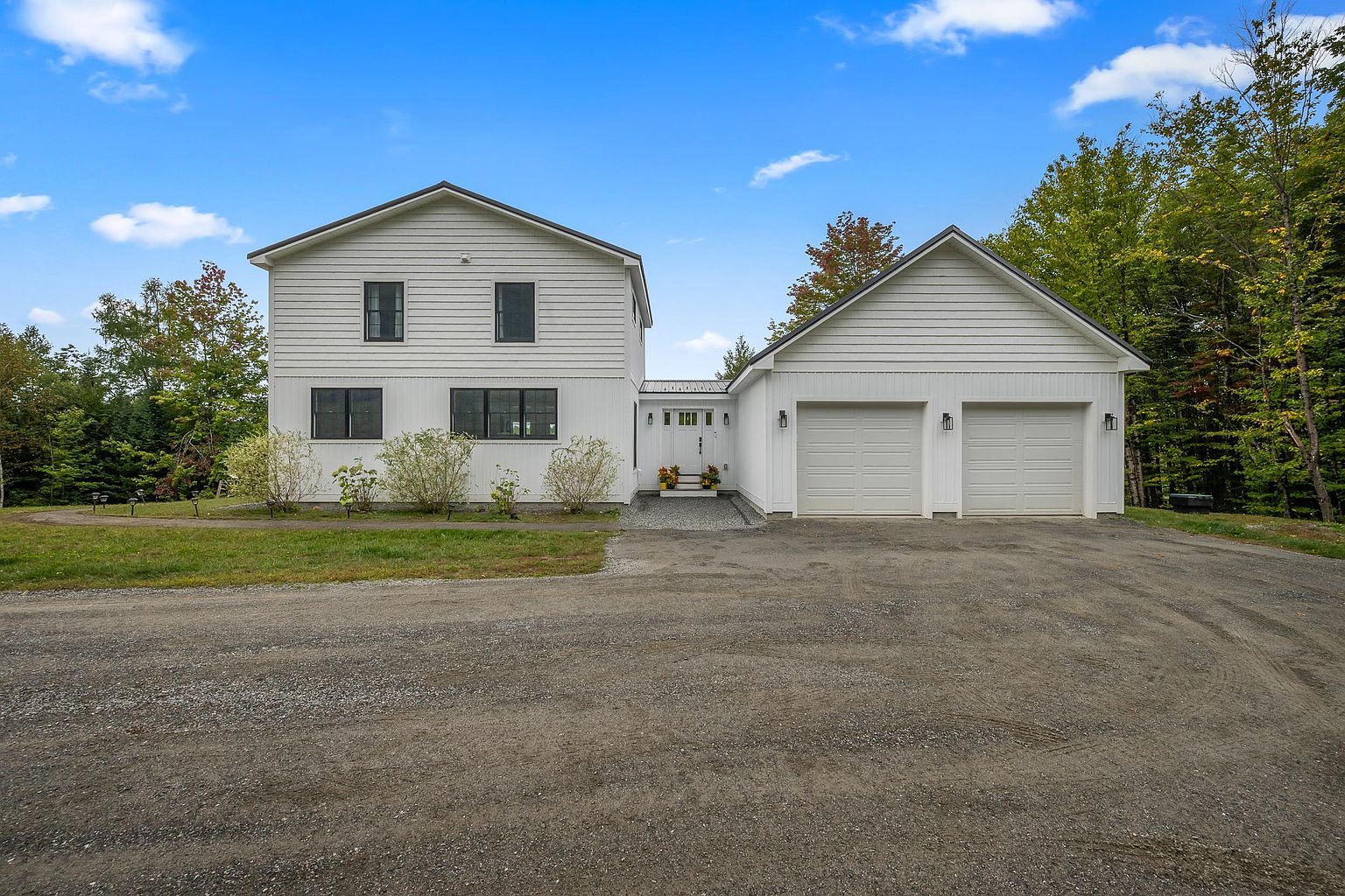 This front-facing view showcases a modern farmhouse-style home with white horizontal siding and a two-car garage connected by a central entryway. The property features dark-framed windows, a gravel driveway, and a natural, wooded backdrop under a bright blue sky. The clean lines and symmetrical design create a welcoming and contemporary curb appeal.