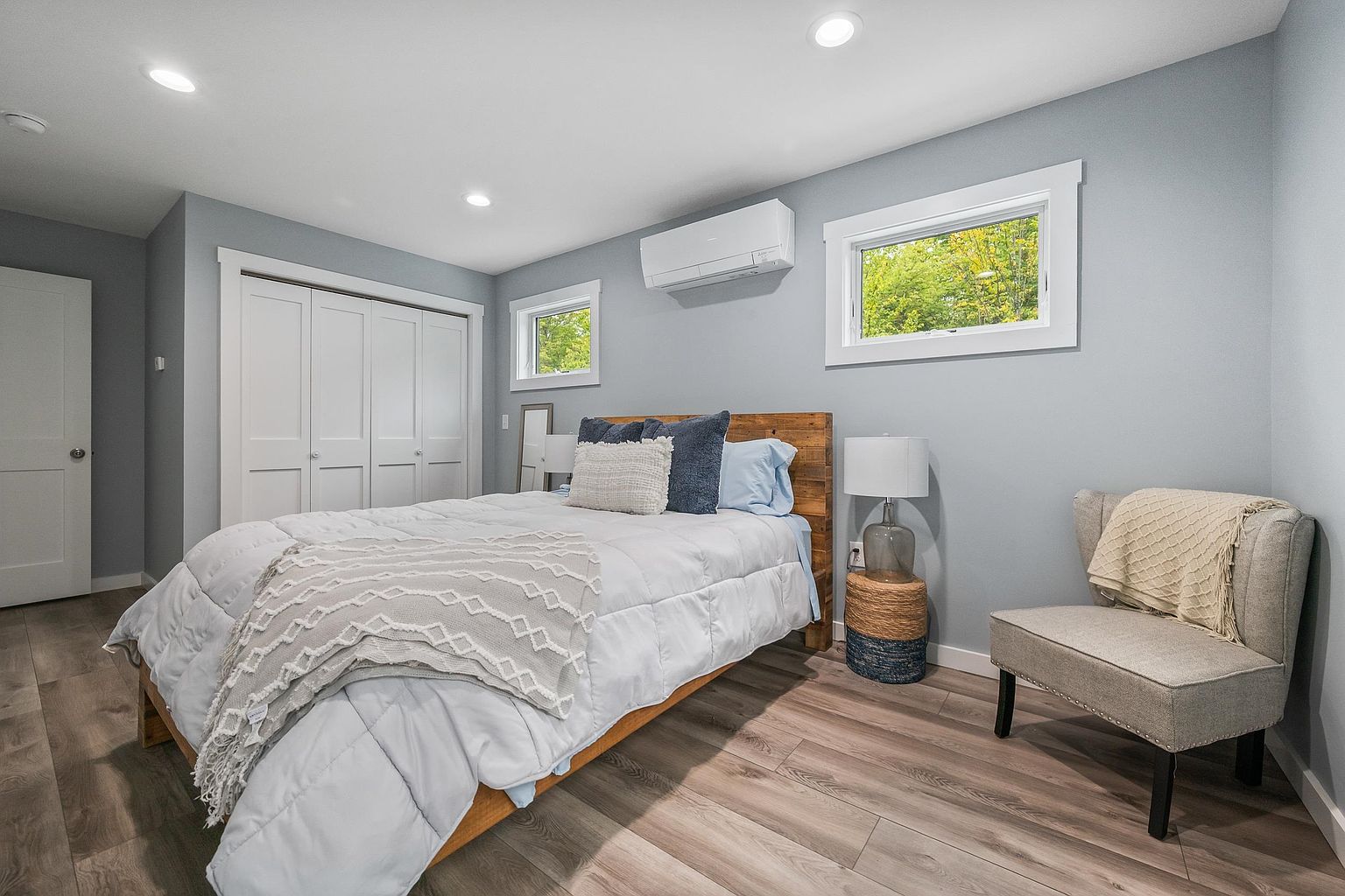 This bright and airy bedroom features light blue walls, wood-look flooring, and a queen-sized bed with a rustic wood headboard. The room is accented by a cozy armchair in the corner, a small woven side table with a lamp, and a wall-mounted ductless mini-split unit for climate control. Two windows provide natural light, while a white closet with folding doors offers storage, creating a clean and inviting atmosphere.
