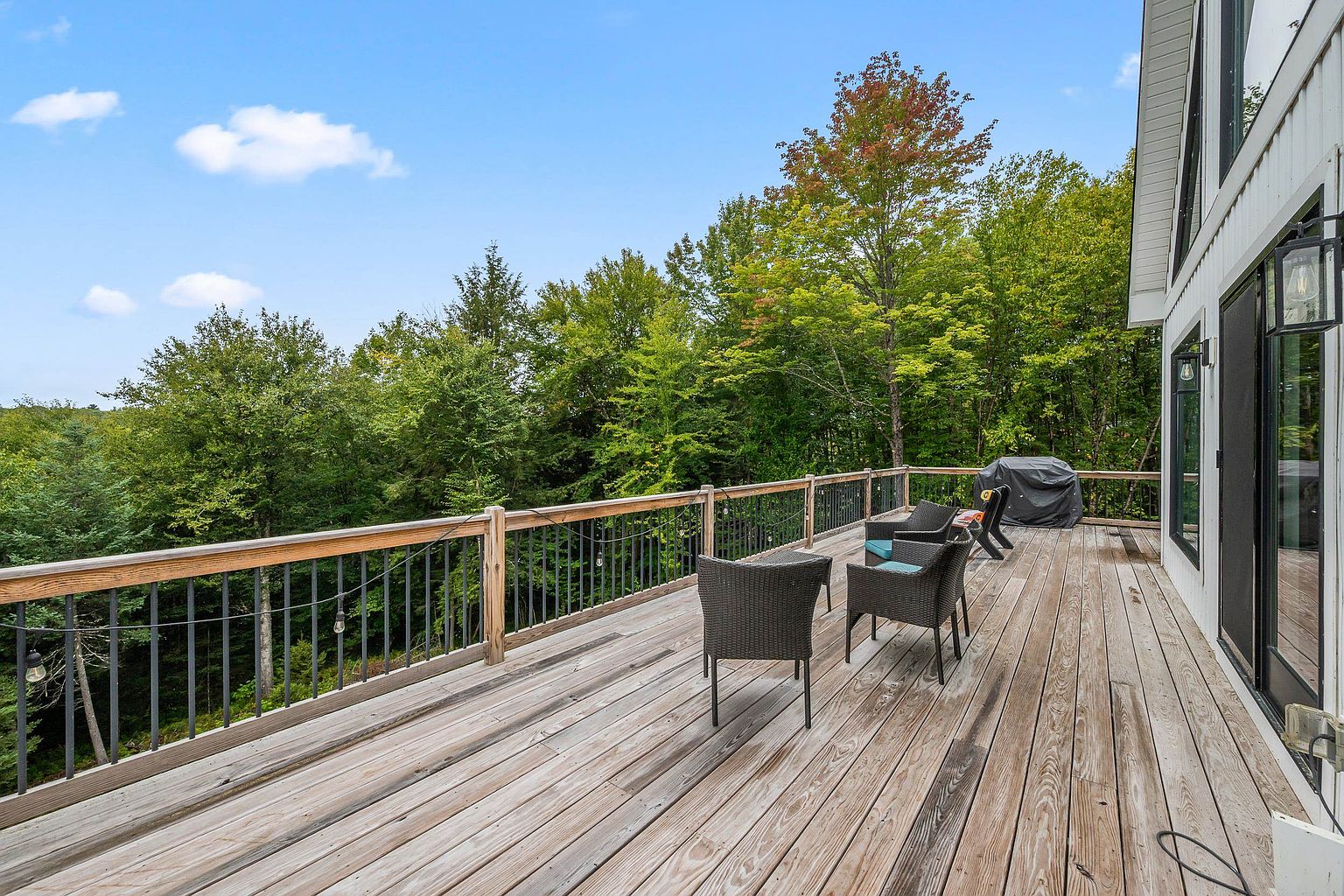 This spacious wooden deck offers a serene outdoor living space with a panoramic view of the surrounding lush forest. The area is furnished with dark wicker chairs and a covered grill, providing a comfortable spot for relaxation or entertaining. The deck features a sturdy railing with metal balusters and is bathed in natural light under a clear blue sky.