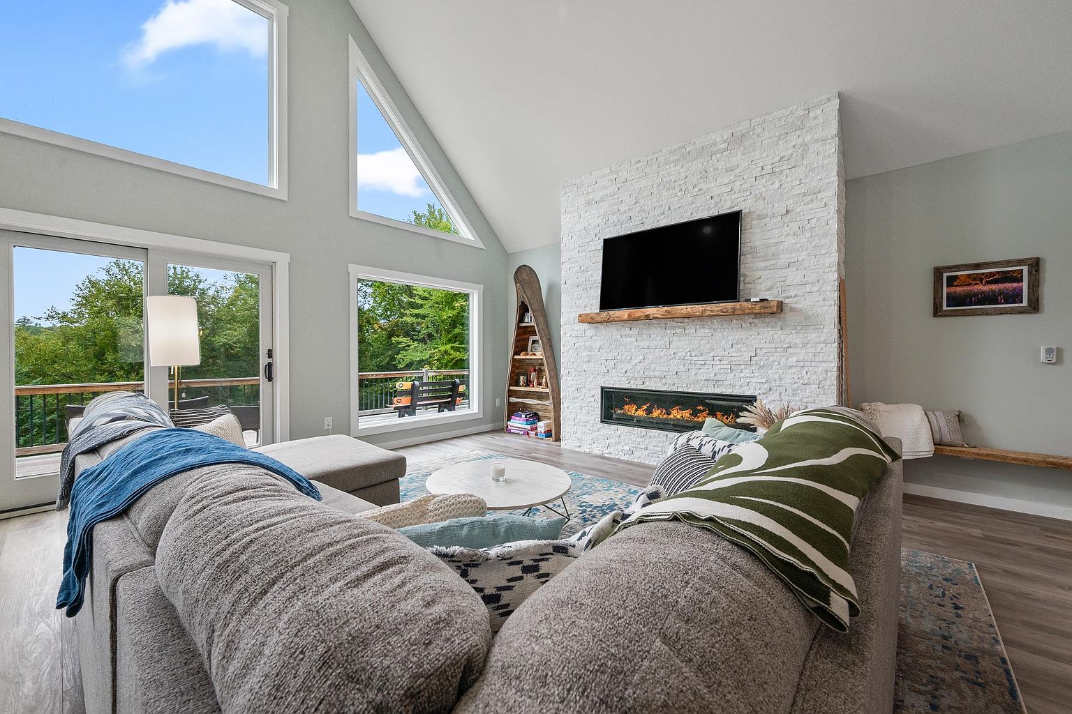 This bright and airy living room features a vaulted ceiling with large windows that flood the space with natural light. A prominent white stacked-stone chimney breast serves as the focal point, housing a linear fireplace and a wall-mounted television above a rustic wood mantel. The room is furnished with a large, comfortable sectional sofa and a circular coffee table, all set against a backdrop of light-toned walls and wood-look flooring.
