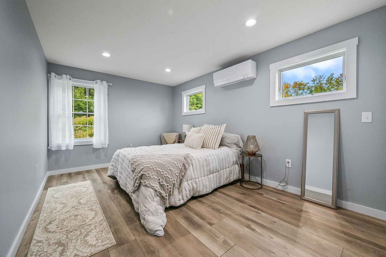 This bright and airy bedroom features light-toned wood-look flooring and soft blue-grey walls, creating a calm and inviting atmosphere. The room is furnished with a comfortable bed dressed in neutral-toned bedding, a small side table with a decorative lamp, and a full-length floor mirror. Natural light streams in through two windows, highlighting the clean, minimalist aesthetic of the space.