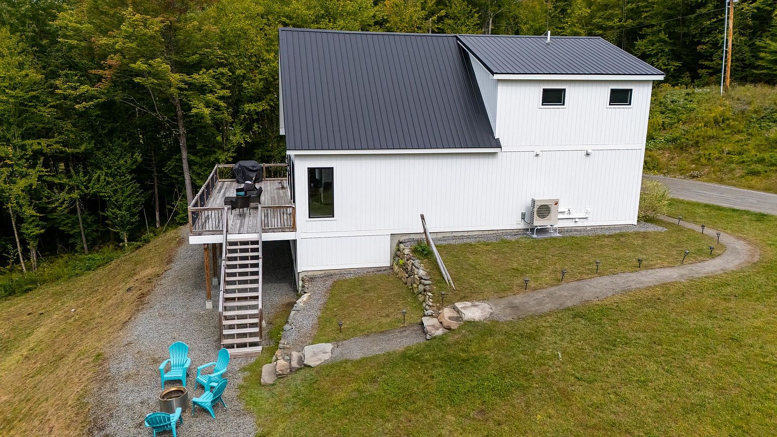 This aerial perspective captures a modern, white-sided home with a dark metal roof, set against a lush, wooded backdrop. The property features a raised wooden deck with stairs leading down to a gravel fire pit area furnished with bright blue chairs, and a stone-lined pathway winds through the grassy yard. The clean lines and minimalist aesthetic of the structure are complemented by the natural, serene landscape surrounding the home.