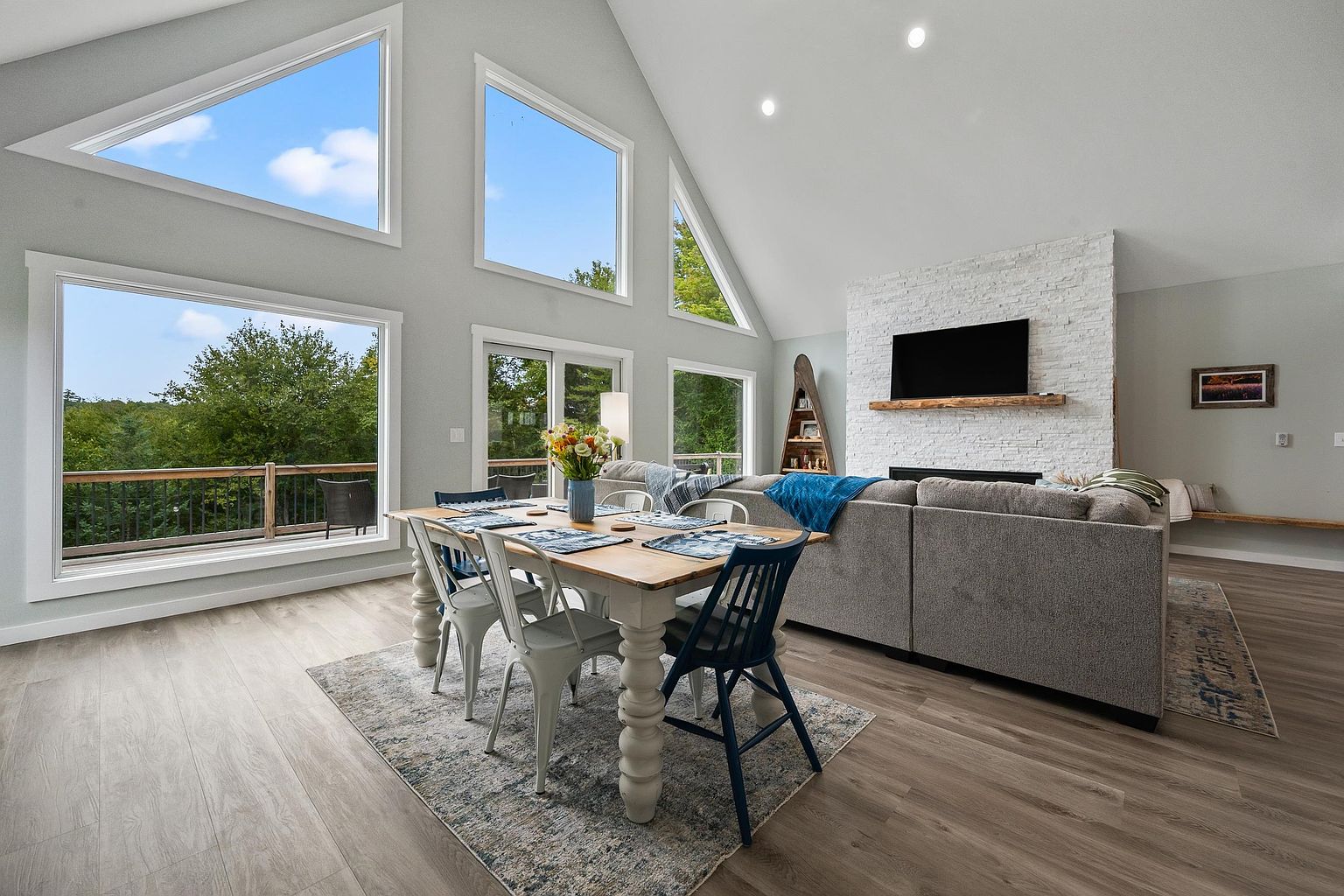 This open-concept living and dining area features a vaulted ceiling and large windows that provide abundant natural light and scenic views. A prominent white stone-clad chimney breast serves as the focal point, complemented by a rustic wood mantel and a large television. The space is furnished with a comfortable gray sectional sofa and a dining table, creating a cohesive and inviting atmosphere for relaxation and entertaining.
