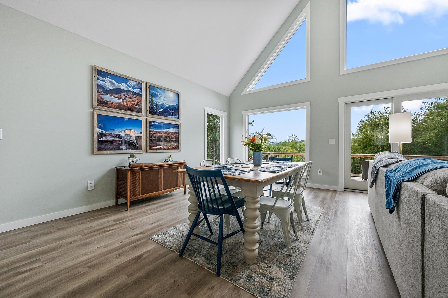 This bright and airy dining area features a rustic wooden table surrounded by a mix of blue and white metal chairs, set atop a patterned area rug. The space is highlighted by a vaulted ceiling and large windows that provide expansive views of the surrounding greenery. A wooden sideboard adorned with framed landscape photography sits against the light-toned wall, creating a welcoming and cohesive aesthetic.