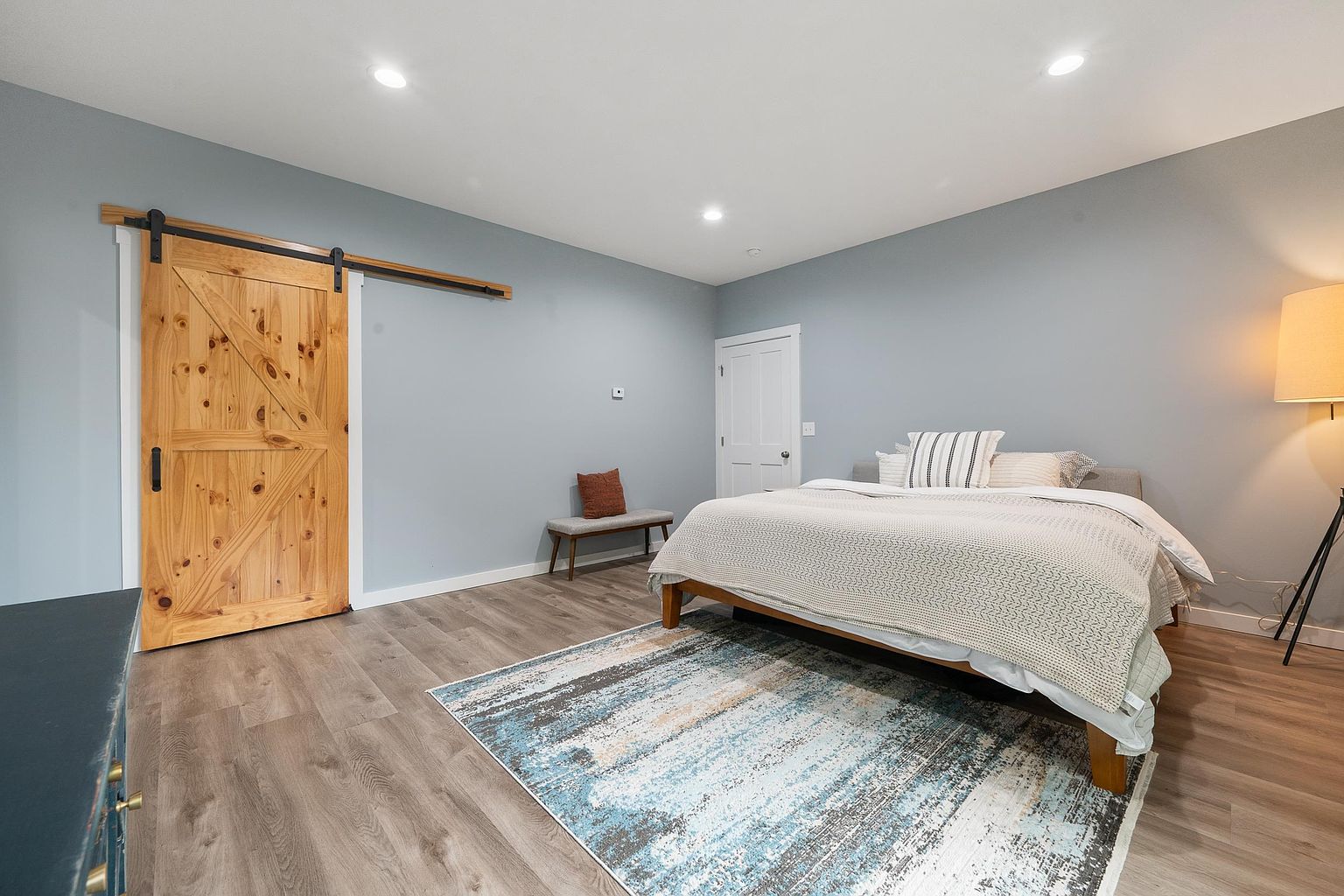This spacious bedroom features light blue walls, wood-look flooring, and a large area rug with a blue and white abstract pattern. A wooden barn-style sliding door adds a rustic touch, while a neutral-toned bed with layered bedding serves as the focal point. The room is illuminated by recessed lighting and a tall floor lamp, creating a calm and inviting atmosphere.