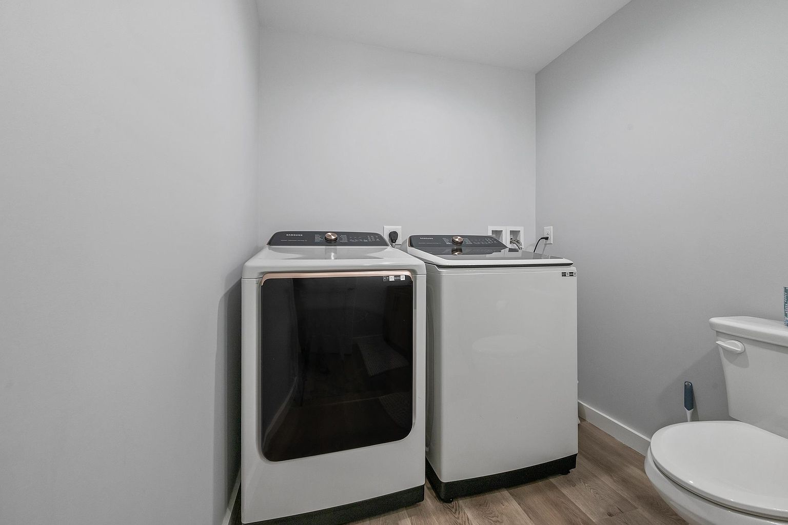 This functional laundry room features a side-by-side Samsung washer and dryer set positioned against a neutral gray wall. To the right, a white toilet is visible, indicating a combined laundry and bathroom space. The room is finished with light wood-look flooring, creating a clean and utilitarian aesthetic.