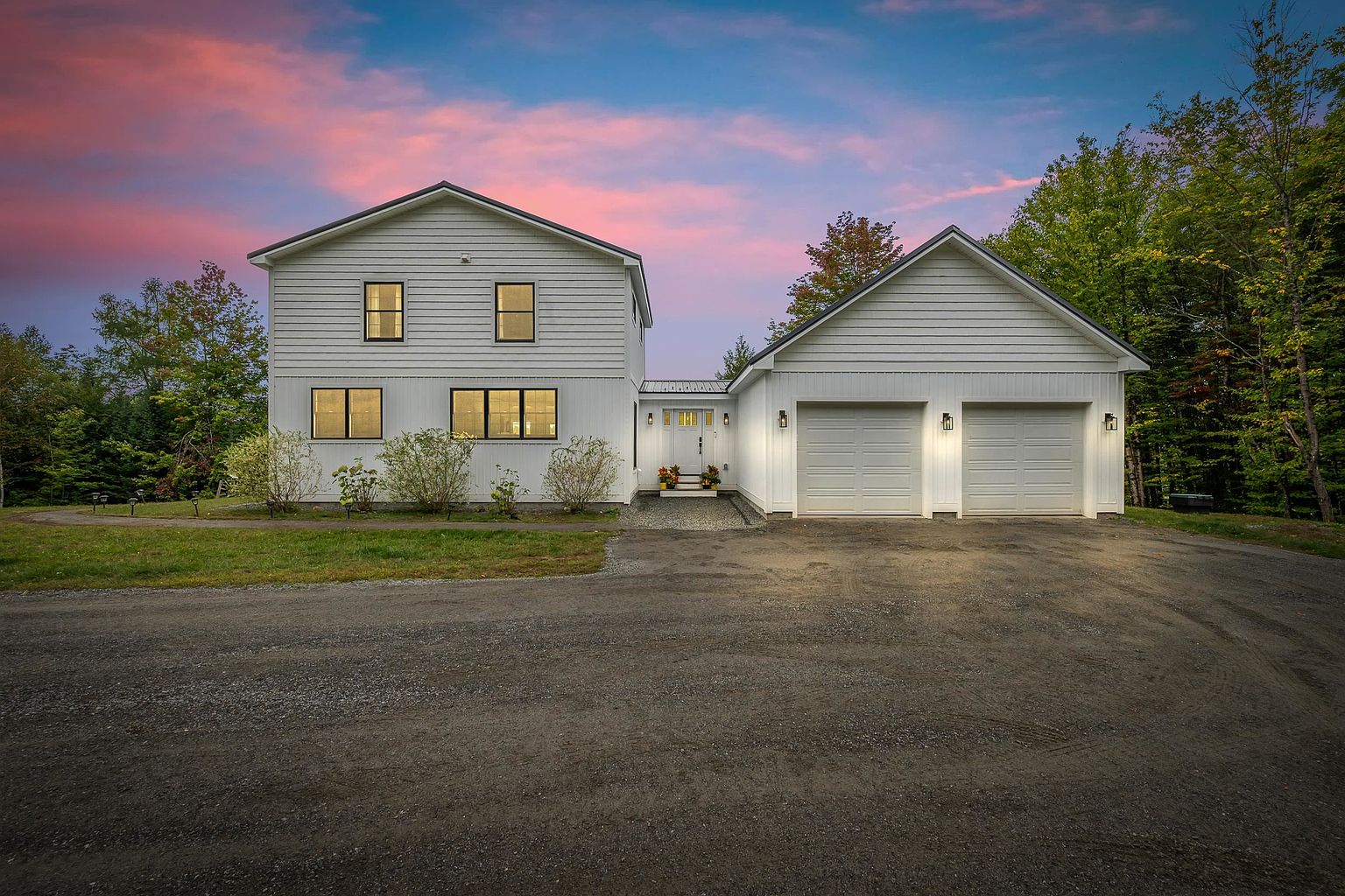 This two-story home features a clean, modern farmhouse aesthetic with white horizontal siding and dark-framed windows. The property includes an attached two-car garage and a central entryway, all set against a vibrant sunset sky. A gravel driveway leads up to the home, which is surrounded by mature trees and a simple, well-maintained lawn.