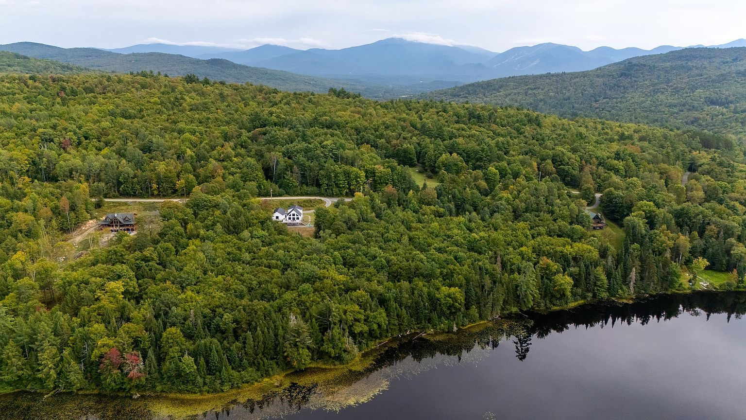 This wide-angle aerial shot captures a secluded residential property nestled within a dense, lush forest bordering a calm lake. The landscape features rolling hills and distant mountain ranges under a cloudy sky, emphasizing the property's private and scenic location. Several homes are visible among the trees, connected by a winding road, showcasing a peaceful, nature-oriented lifestyle.