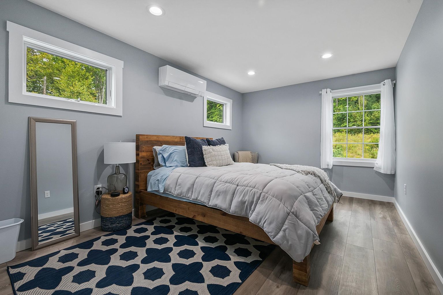 This bright and airy bedroom features light blue walls, wood-look flooring, and a rustic wooden bed frame topped with a grey comforter. A patterned navy and white area rug anchors the space, while a full-length mirror and a small side table with a lamp add functional charm. Large windows on two walls provide ample natural light and views of the surrounding greenery, creating a peaceful and inviting atmosphere.