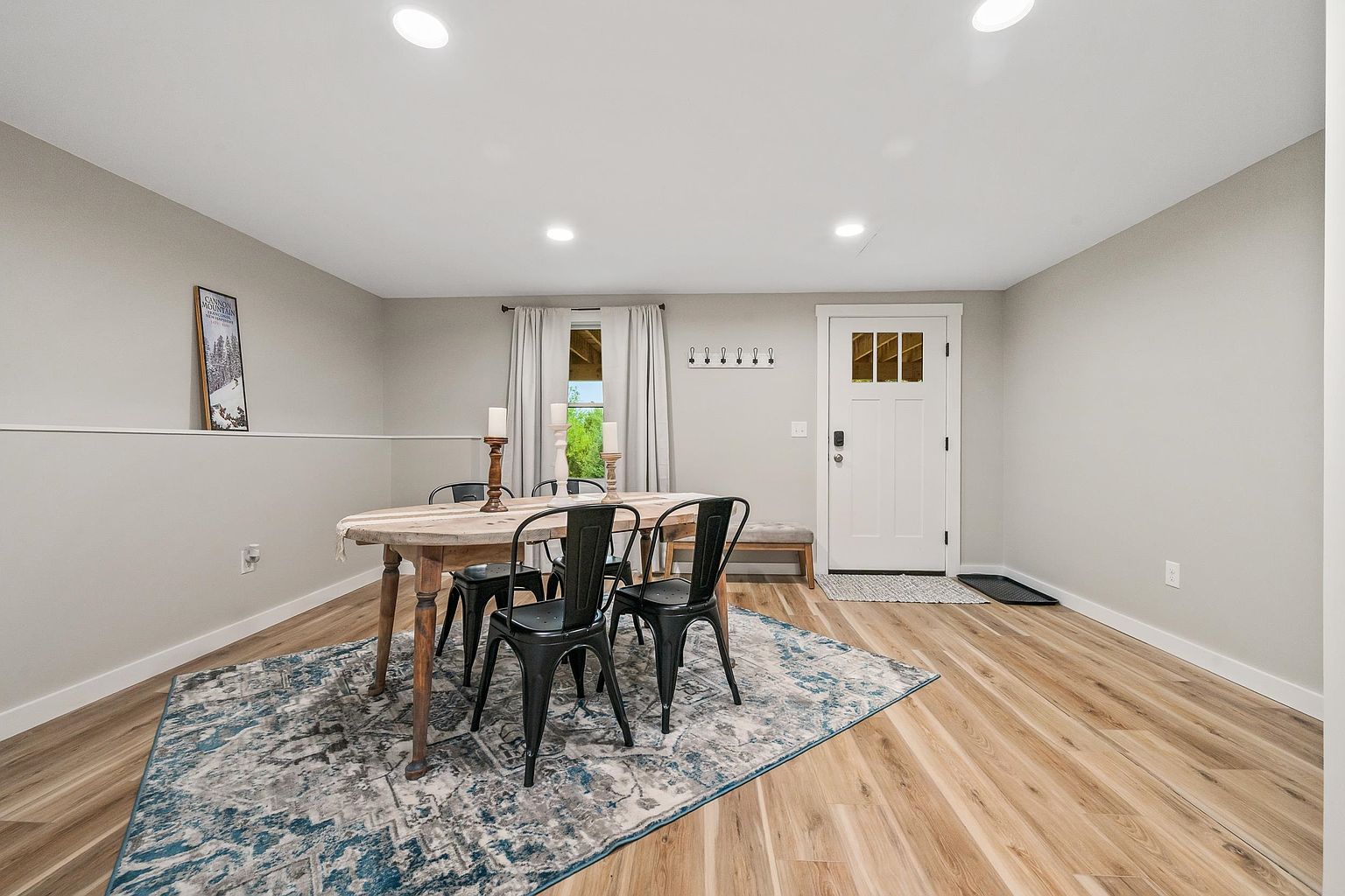 This dining room features a rustic wooden table surrounded by four black metal chairs, set atop a large, patterned blue and grey area rug. The space is illuminated by recessed lighting and includes a white front door with glass panes, a wall-mounted coat rack, and a bench tucked against the wall. The neutral wall color and light wood-look flooring create a bright, welcoming atmosphere.