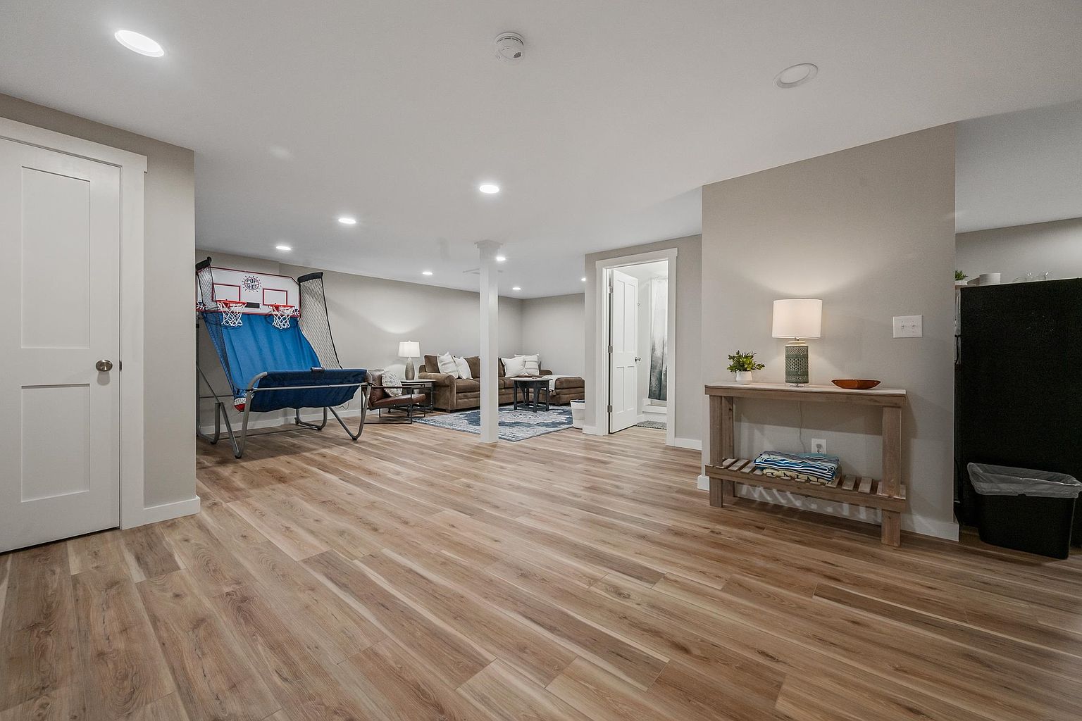 This spacious, finished basement features light wood-look flooring and neutral walls, creating a versatile living area. The room includes a dedicated lounge space with a brown sectional sofa and a blue area rug, alongside a recreational area equipped with a dual-hoop basketball arcade game. A console table with a lamp provides a welcoming accent near the entrance, while recessed lighting throughout ensures a bright and inviting atmosphere.