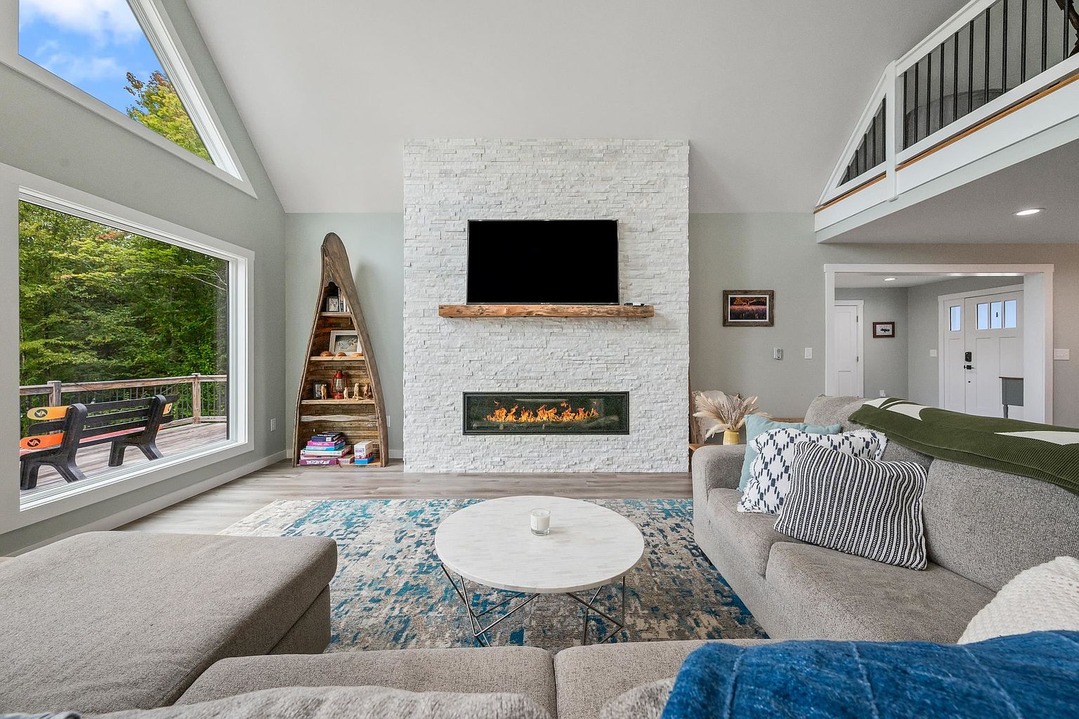 This bright and airy living room features a vaulted ceiling and a prominent stone-clad chimney breast housing a modern linear fireplace and a wall-mounted television. A unique boat-shaped shelving unit stands to the left, while a large window provides a scenic view of the outdoor deck. The space is anchored by a plush gray sectional sofa and a round coffee table, creating a comfortable and inviting atmosphere.
