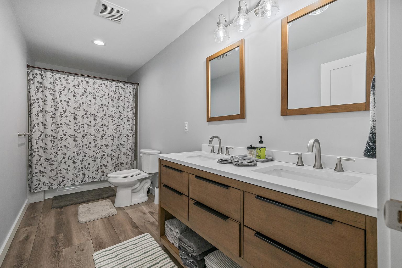 This modern bathroom features a double-sink vanity with a light wood finish, dark hardware, and a white countertop. The space includes a shower area with a patterned curtain, a toilet, and two separate mirrors mounted above the vanity. The room is finished with light gray walls and wood-look flooring, creating a clean and contemporary aesthetic.