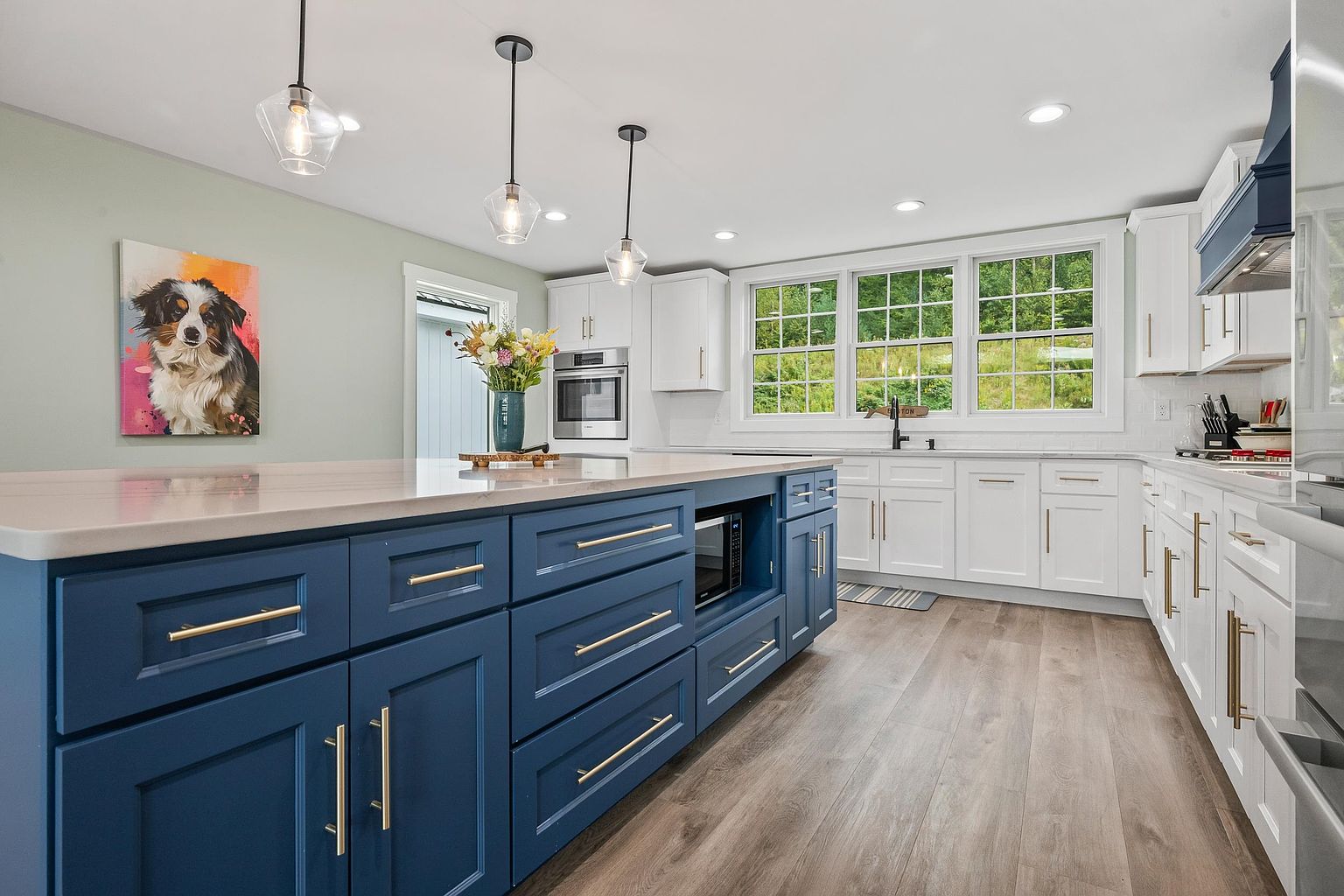 This bright, modern kitchen features a prominent navy blue center island with gold-toned hardware and a built-in microwave. The perimeter is lined with crisp white cabinetry, light-colored countertops, and a subway tile backsplash, all illuminated by three glass pendant lights. Large windows above the sink provide a clear view of the outdoors, while a colorful dog portrait adds a personal touch to the light green wall.