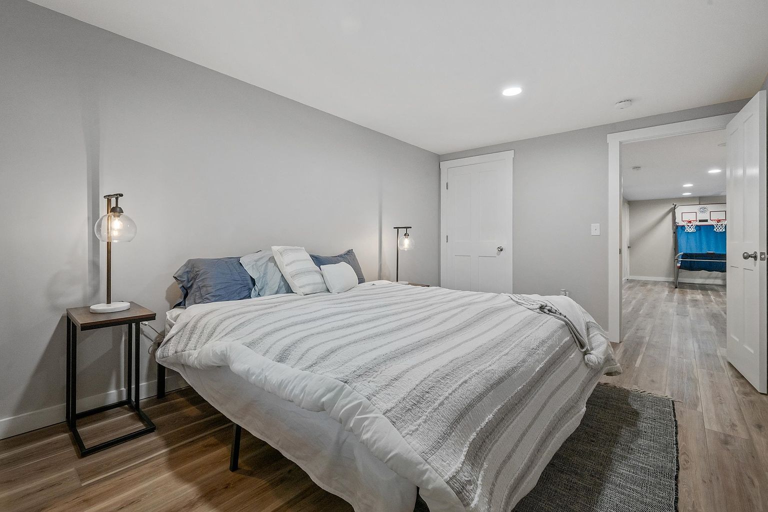This bedroom features a neutral color palette with light gray walls and wood-look flooring, creating a calm and inviting atmosphere. A bed with a striped comforter is centered against the wall, flanked by two minimalist bedside tables with modern glass globe lamps. An open doorway leads to an adjacent room, providing a glimpse of a recreational space with a basketball arcade game, suggesting a versatile layout.
