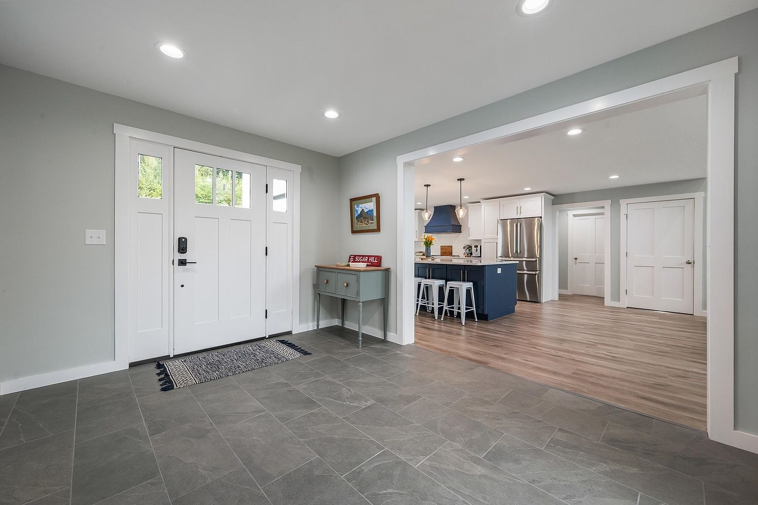 This bright entryway features large-format gray tile flooring that transitions into light wood-toned flooring leading into the adjacent kitchen. A white front door with glass panels provides natural light, while a console table with a decorative sign adds a welcoming touch. The open-concept layout offers a clear view into the kitchen, which is highlighted by a deep blue island, white cabinetry, and modern pendant lighting.