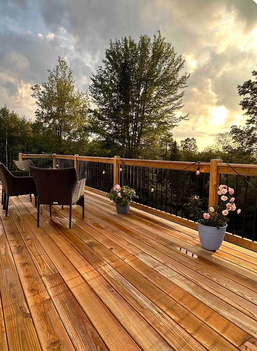 This elevated wooden deck offers a serene outdoor living space with a clear view of a lush, forested landscape under a dramatic sunset sky. The deck features dark wicker seating, potted flowering plants, and string lights draped along the wooden railing, creating an inviting atmosphere for relaxation. The warm, golden light of the setting sun highlights the natural wood grain of the decking, emphasizing the peaceful and scenic nature of the property.
