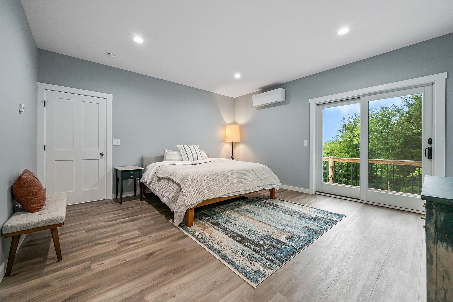 This bright and airy primary bedroom features light-toned wood-look flooring and soft blue-grey walls, creating a serene atmosphere. A central bed with a neutral-toned quilt is flanked by a minimalist nightstand and a floor lamp, while a small bench sits near the door. Large sliding glass doors provide access to an outdoor deck, allowing for ample natural light and a scenic view of the surrounding trees.