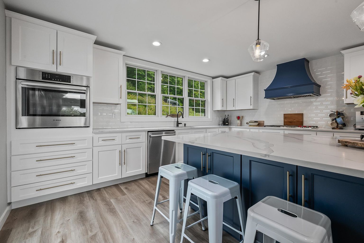 This bright, modern kitchen features a striking contrast between white perimeter cabinetry and a deep blue central island topped with a light-toned stone countertop. The space is equipped with stainless steel appliances, including a built-in wall oven and a matching blue range hood, while light-toned wood-look flooring adds warmth. Large windows above the sink provide a clear view of the lush green exterior, and a single glass pendant light hangs over the island, creating a clean and inviting atmosphere.