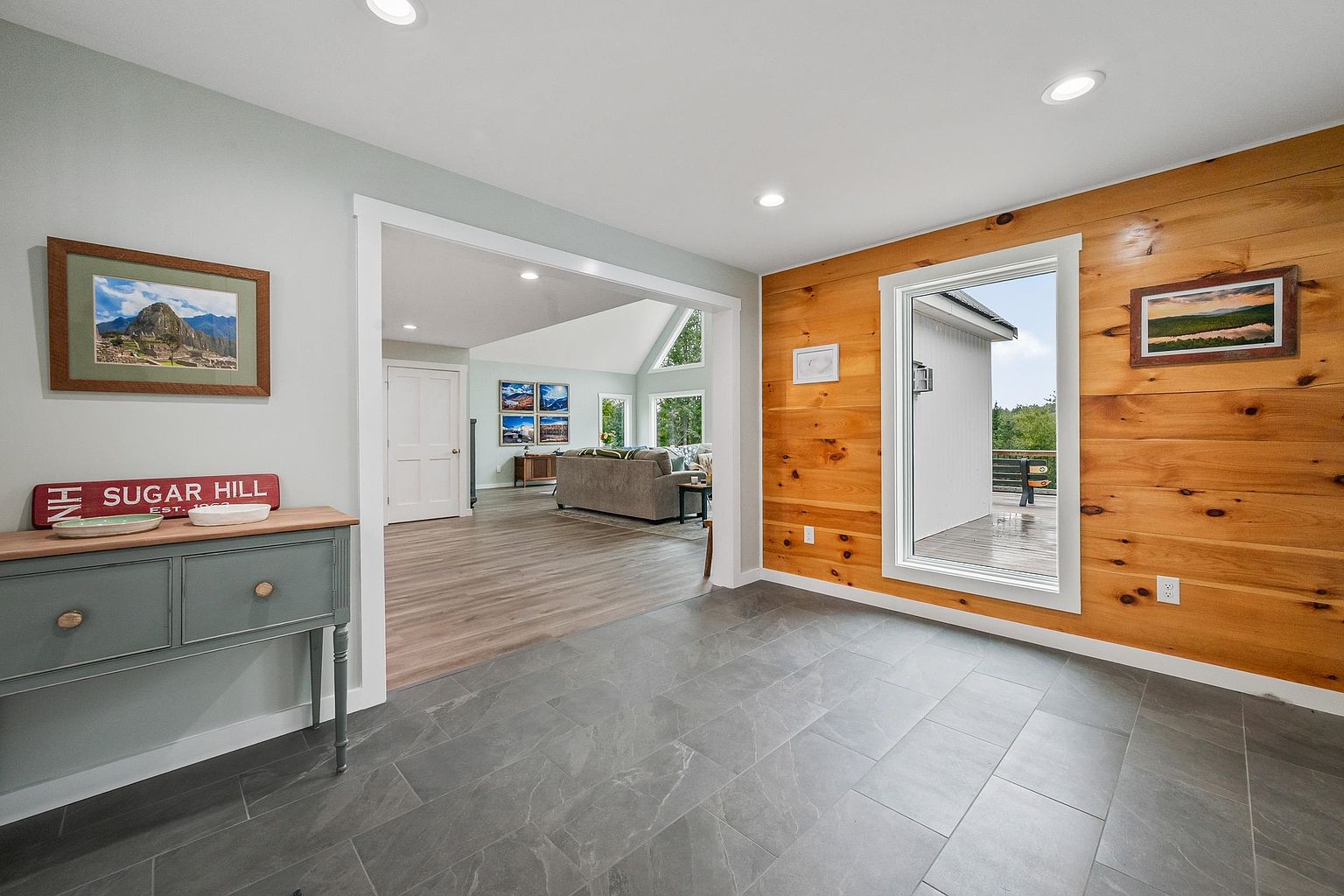 This bright entryway features large-format gray tile flooring that transitions into light-toned wood flooring in the adjacent living area. A prominent wood-paneled accent wall adds warmth to the space, while a large window provides a clear view of the outdoor deck. The open-concept layout creates a seamless flow between the foyer and the main living space, which is visible through a wide, white-trimmed doorway.