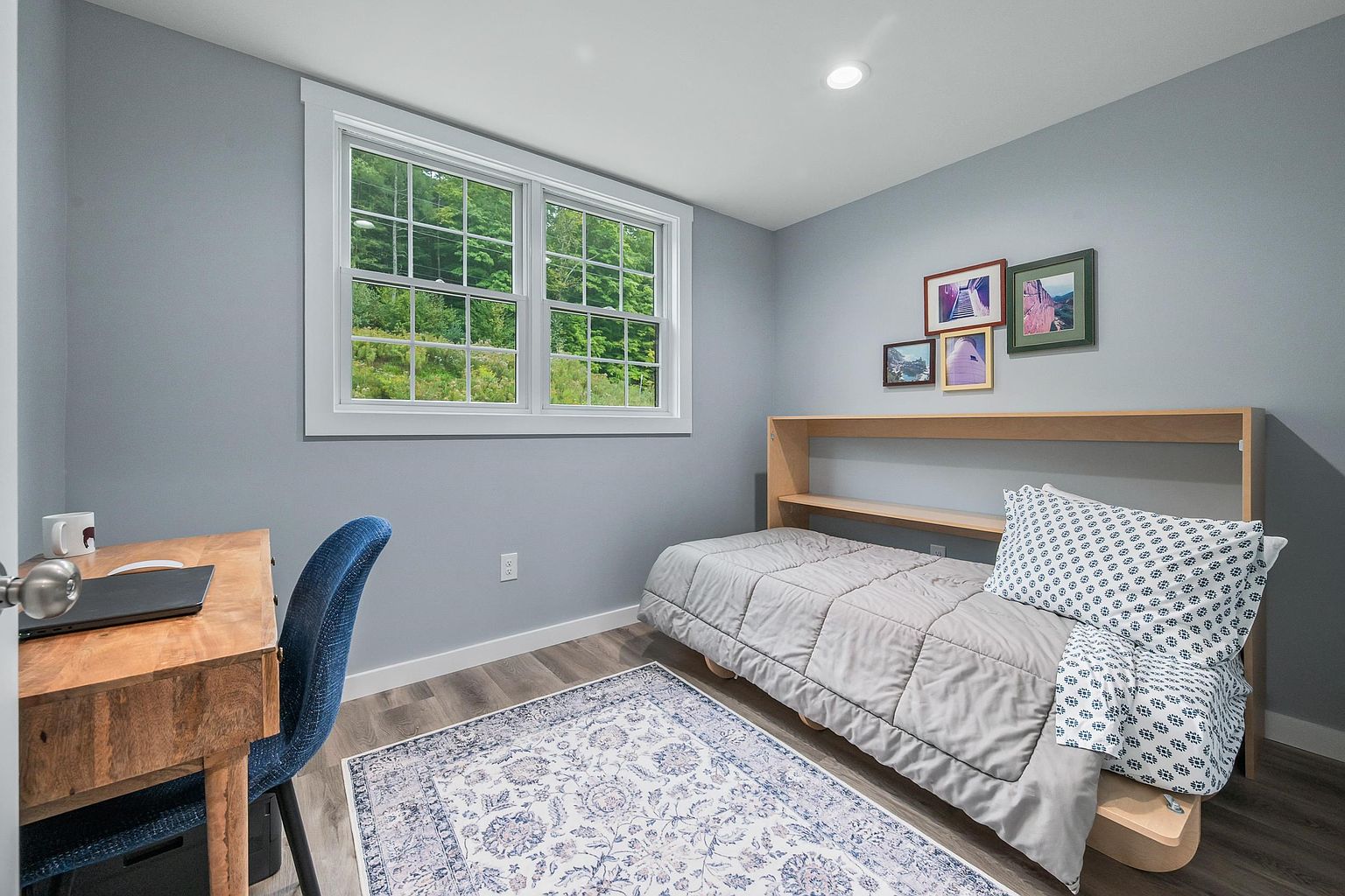 This cozy bedroom features light blue walls, wood-look flooring, and a patterned area rug that adds texture to the space. A twin bed with a grey comforter and a patterned pillow is positioned against a light wood headboard with integrated shelving, while a wooden desk and blue chair provide a functional workspace. A large window with a white frame allows natural light to fill the room, offering a view of the lush green trees outside.