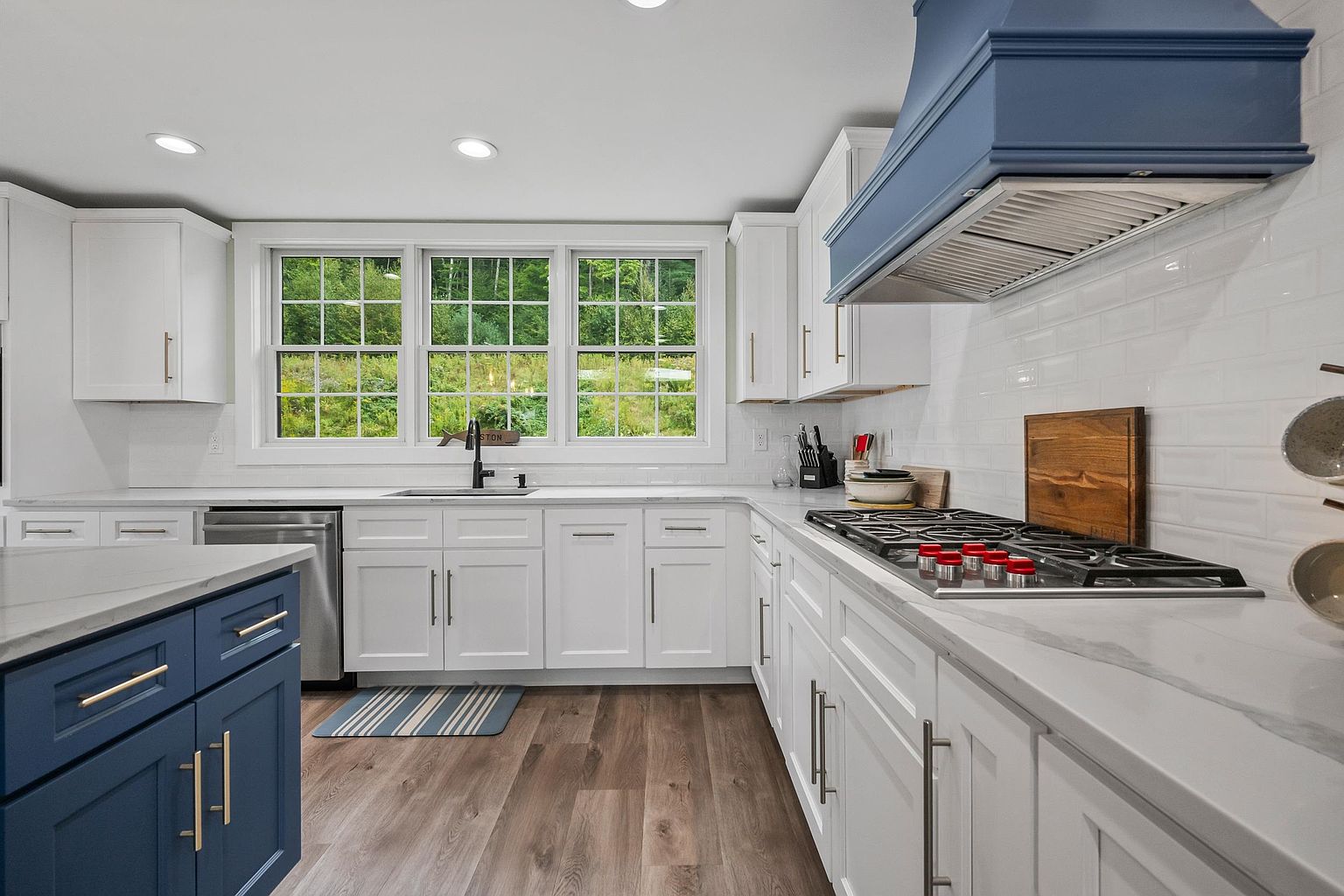 This bright, modern kitchen features a striking contrast between white perimeter cabinetry and a deep blue island and range hood. The space is equipped with stainless steel appliances, a gas cooktop with red knobs, and light-toned wood-look flooring. Large windows above the sink provide a clear view of the lush green landscape outside, creating an airy and inviting atmosphere.