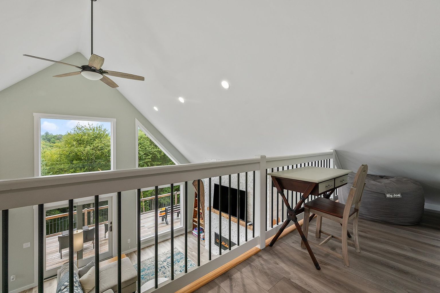 This lofted space overlooks the main living area, featuring a vaulted ceiling and a wooden railing that provides a clear view of the room below. The area is furnished with a small writing desk and a beanbag chair, creating a cozy nook on the wood-look flooring. Large windows allow natural light to fill the space, highlighting the airy, open-concept design of the home.