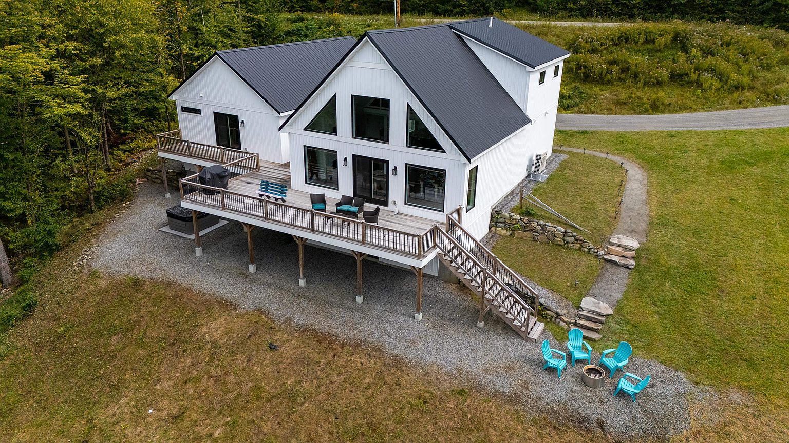 This aerial perspective captures a modern farmhouse-style home with white vertical siding and a dark metal roof, set against a backdrop of dense forest. A spacious wooden deck wraps around the rear, featuring a hot tub, outdoor seating, and a grill, while a gravel path leads to a fire pit area with bright blue Adirondack chairs. The home's design emphasizes clean lines and large windows, creating a bright and inviting atmosphere in a serene, rural setting.